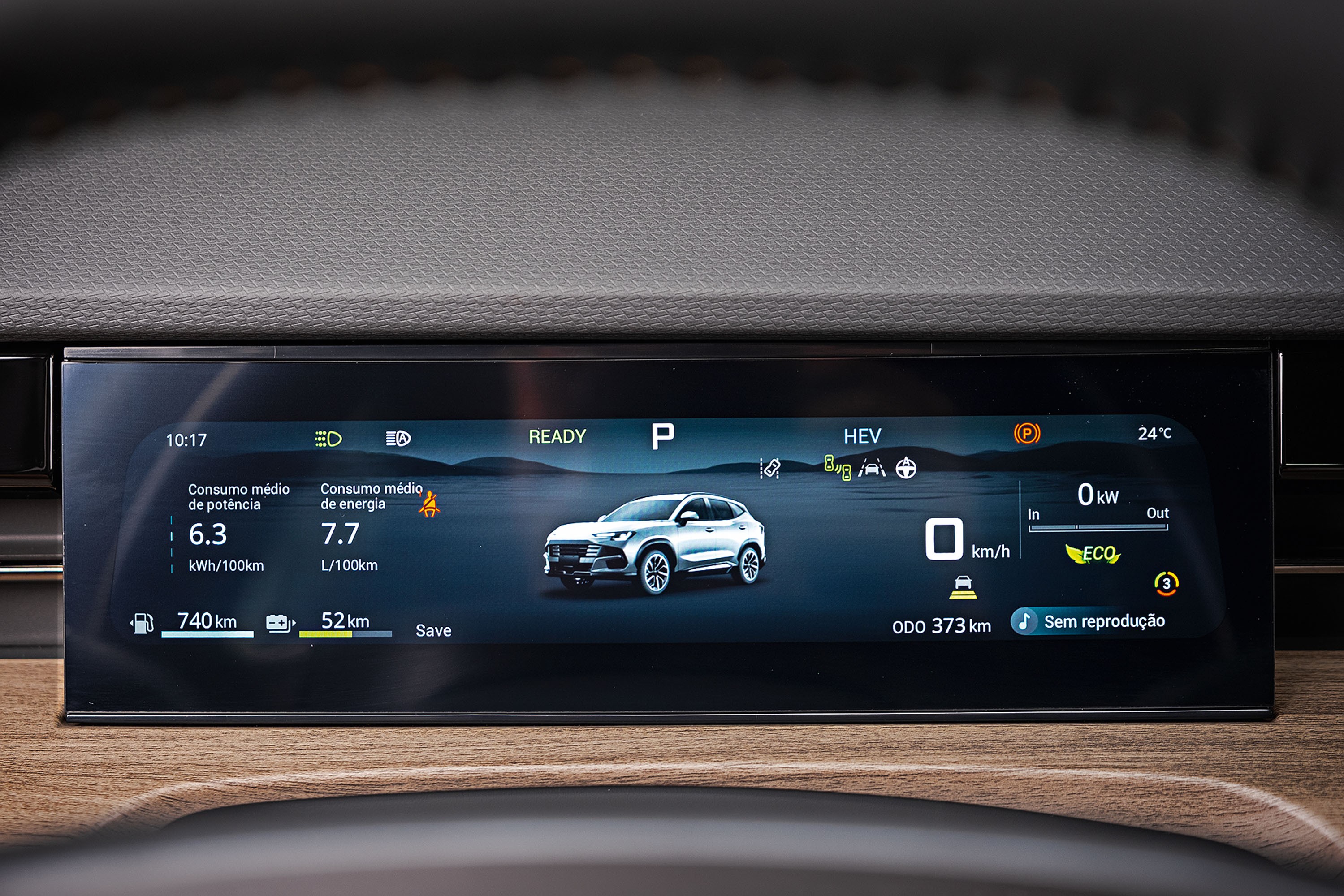 Painel digital de carro exibindo informa&ccedil;&otilde;es de consumo, autonomia e um SUV branco ao centro. O consumo de pot&ecirc;ncia &eacute; 6.3 kWh/100km e o de energia 7.7 L/100km. A autonomia &eacute; de 740 km de combust&iacute;vel e 52 km de bateria. O carro est&aacute; em modo P (estacionado) e HEV (h&iacute;brido), com temperatura de 24&deg;C. O od&ocirc;metro marca 000 373 km e n&atilde;o h&aacute; reprodu&ccedil;&atilde;o de m&iacute;dia. O fundo &eacute; uma paisagem montanhosa azul escura