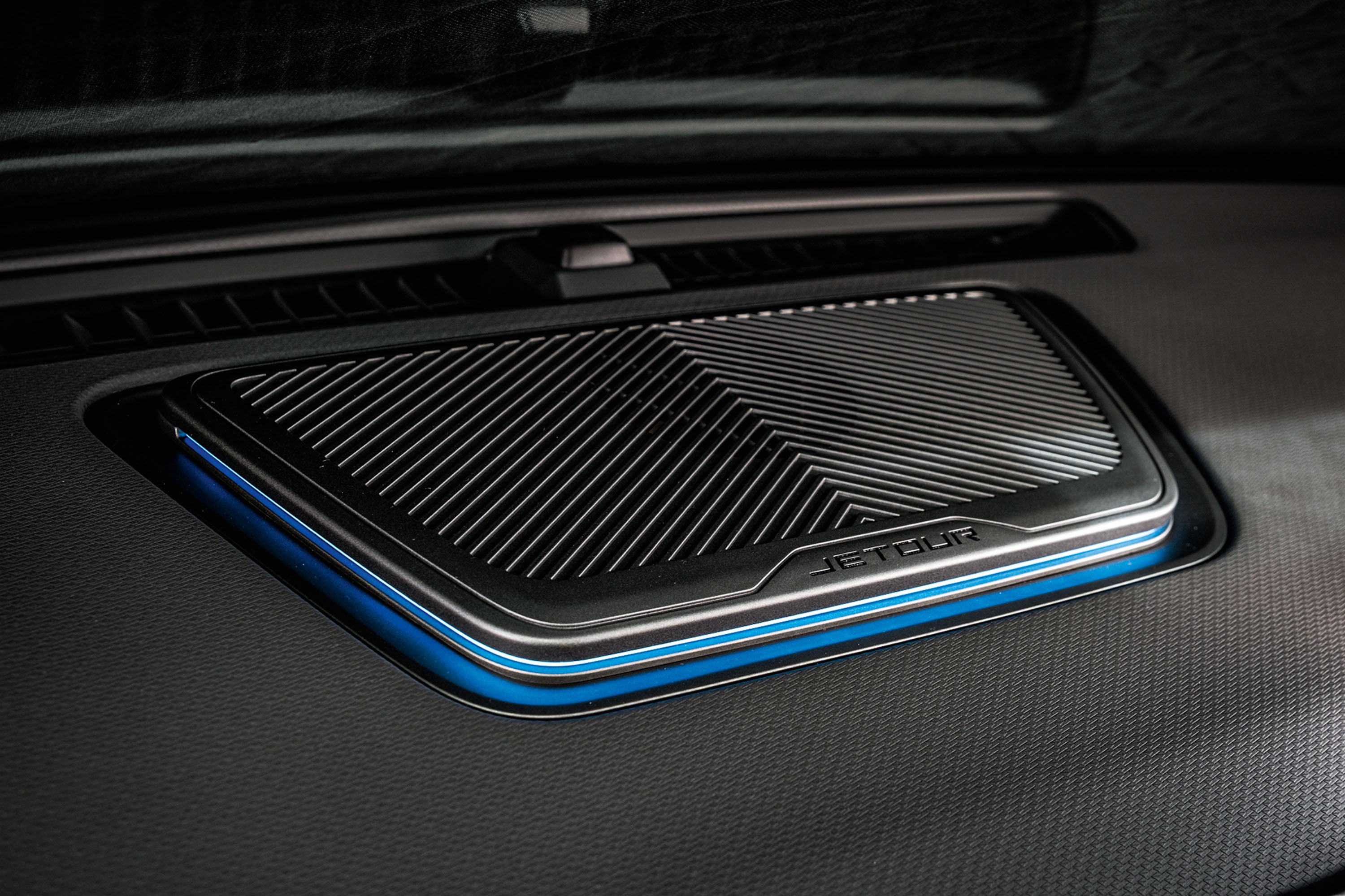 Detalhe do interior de um carro, mostrando um alto-falante retangular com grade met&aacute;lica e ilumina&ccedil;&atilde;o ambiente azul ao redor, sobre um painel escuro texturizado. A marca JETOUR est&aacute; gravada na borda do alto-falante