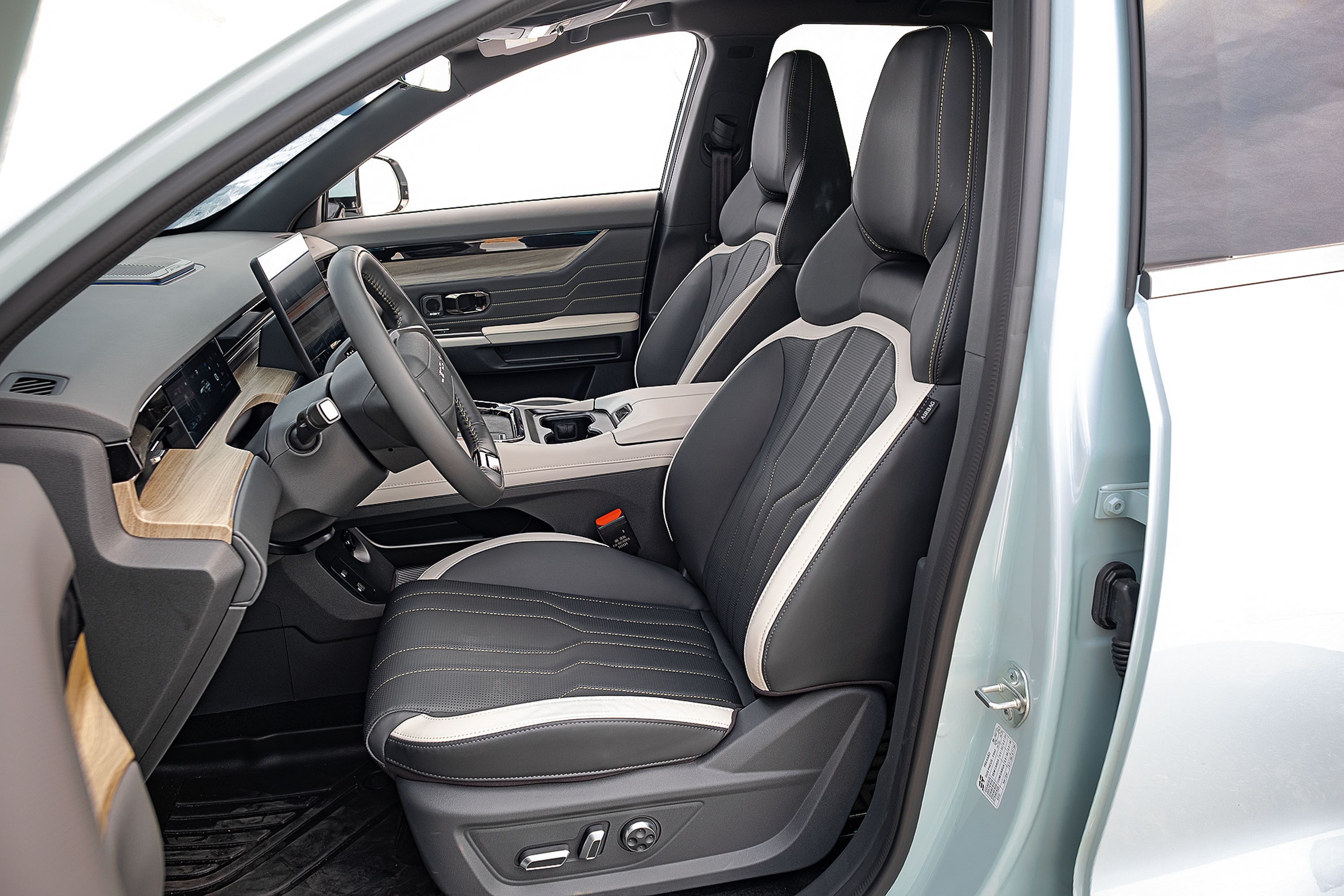 Interior de um carro moderno, com bancos esportivos em couro preto e detalhes brancos, costura aparente. O painel tem acabamento em madeira clara e uma tela digital. O volante &eacute; multifuncional, e o console central possui porta-copos. A porta do motorista est&aacute; aberta, revelando a cor azul-clara da lataria. O ambiente &eacute; clean e sofisticado