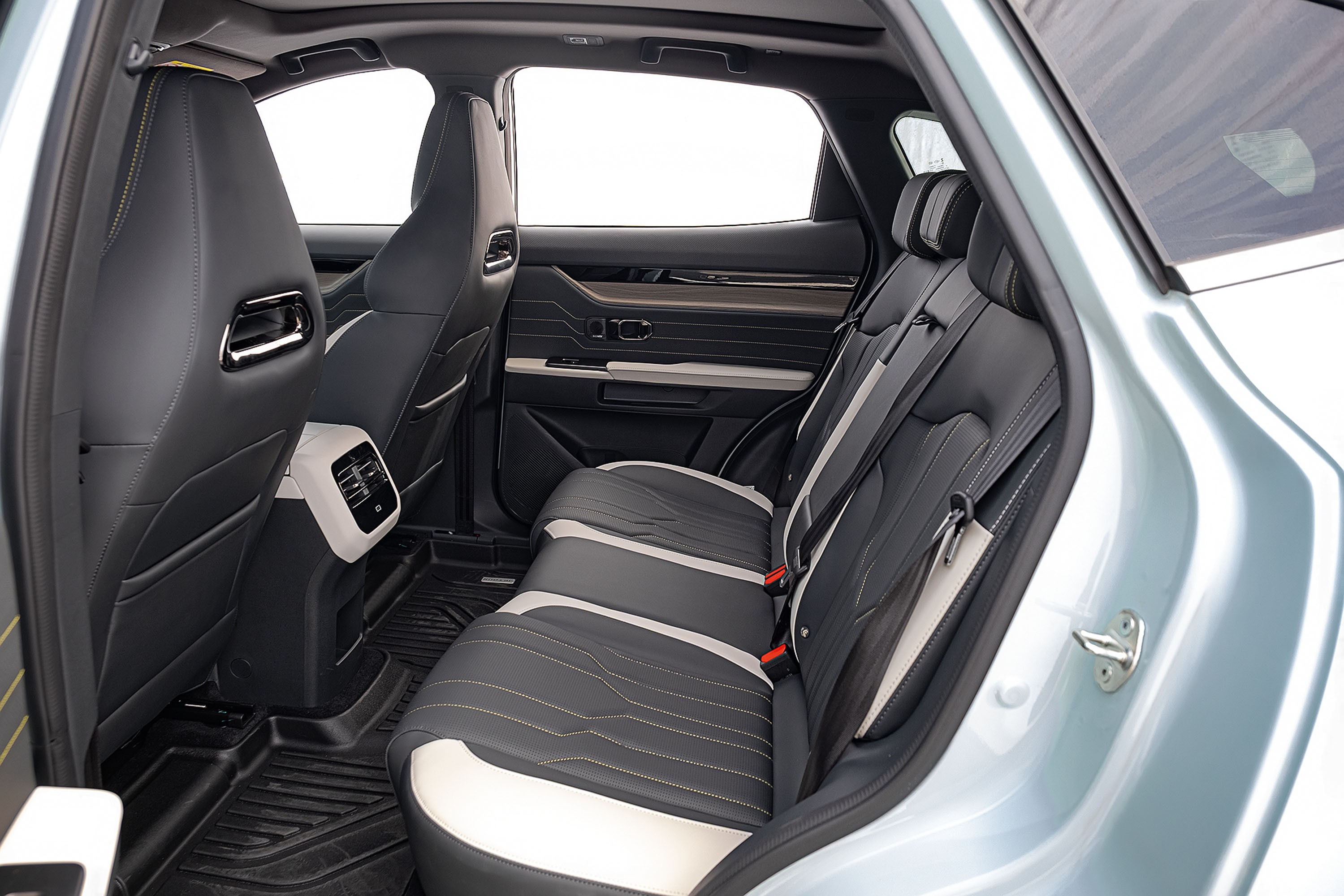Interior de um carro com bancos traseiros em couro preto e detalhes brancos, cintos de seguran&ccedil;a cinzas e tapetes pretos. As janelas est&atilde;o abertas, mostrando o exterior claro. No centro, sa&iacute;das de ar e controles de temperatura.