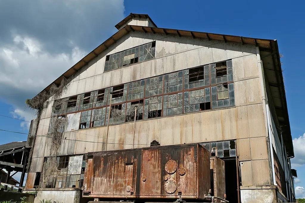 Antiga f&aacute;brica abandonada com paredes de chapa met&aacute;lica, muitas janelas quebradas e vegeta&ccedil;&atilde;o seca subindo pela lateral. Um grande tanque de metal enferrujado est&aacute; em primeiro plano, sob um c&eacute;u azul com nuvens brancas