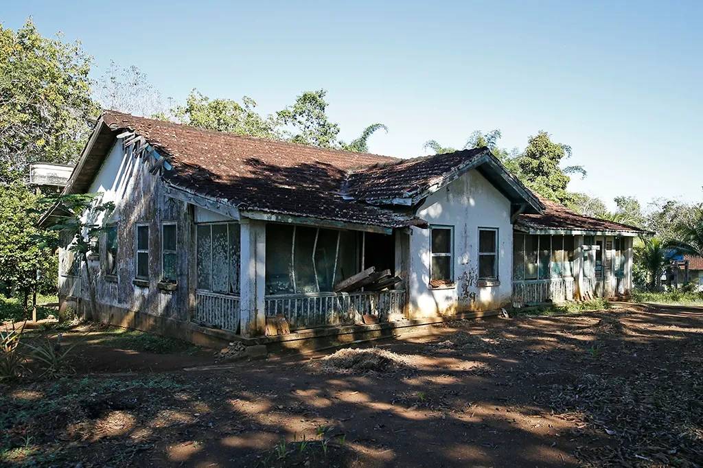 Casa antiga e abandonada, com paredes descascadas, telhado de telhas coloniais marrons e varanda com grades brancas e janelas quebradas. O terreno ao redor &eacute; de terra batida com vegeta&ccedil;&atilde;o rasteira e &aacute;rvores ao fundo, sob c&eacute;u azul claro