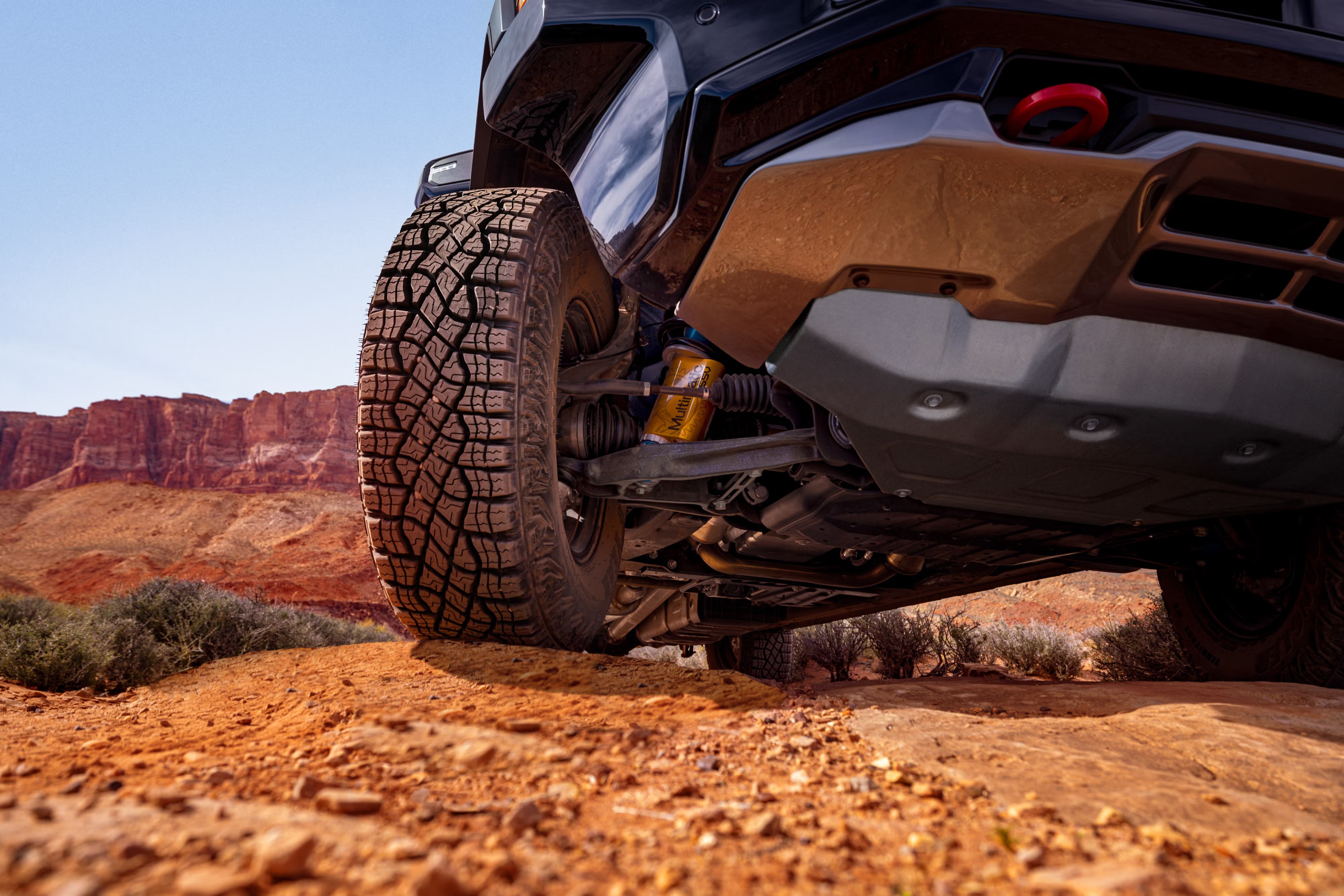 Pneu robusto de um ve&iacute;culo off-road em primeiro plano, sobre terra avermelhada e pedras. Ao fundo, montanhas rochosas e c&eacute;u azul claro. A suspens&atilde;o amarela e a parte inferior do carro s&atilde;o vis&iacute;veis, indicando preparo para terrenos acidentados
