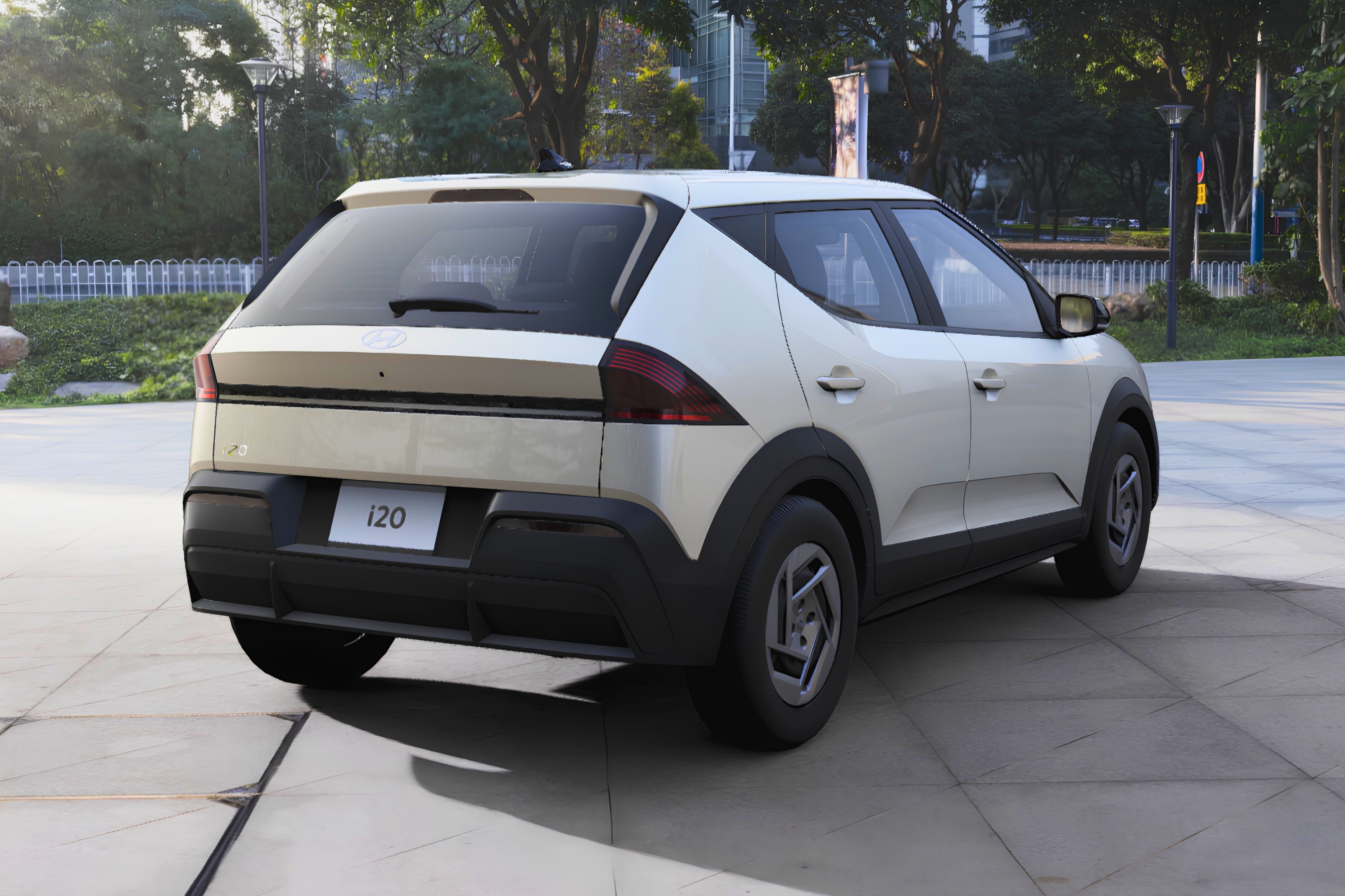 Carro branco Hyundai i20 estacionado em uma &aacute;rea pavimentada, visto de tr&aacute;s e ligeiramente &agrave; direita. O ve&iacute;culo tem detalhes em preto na parte inferior e rodas prateadas. Ao fundo, &aacute;rvores e edif&iacute;cios borrados sob luz solar