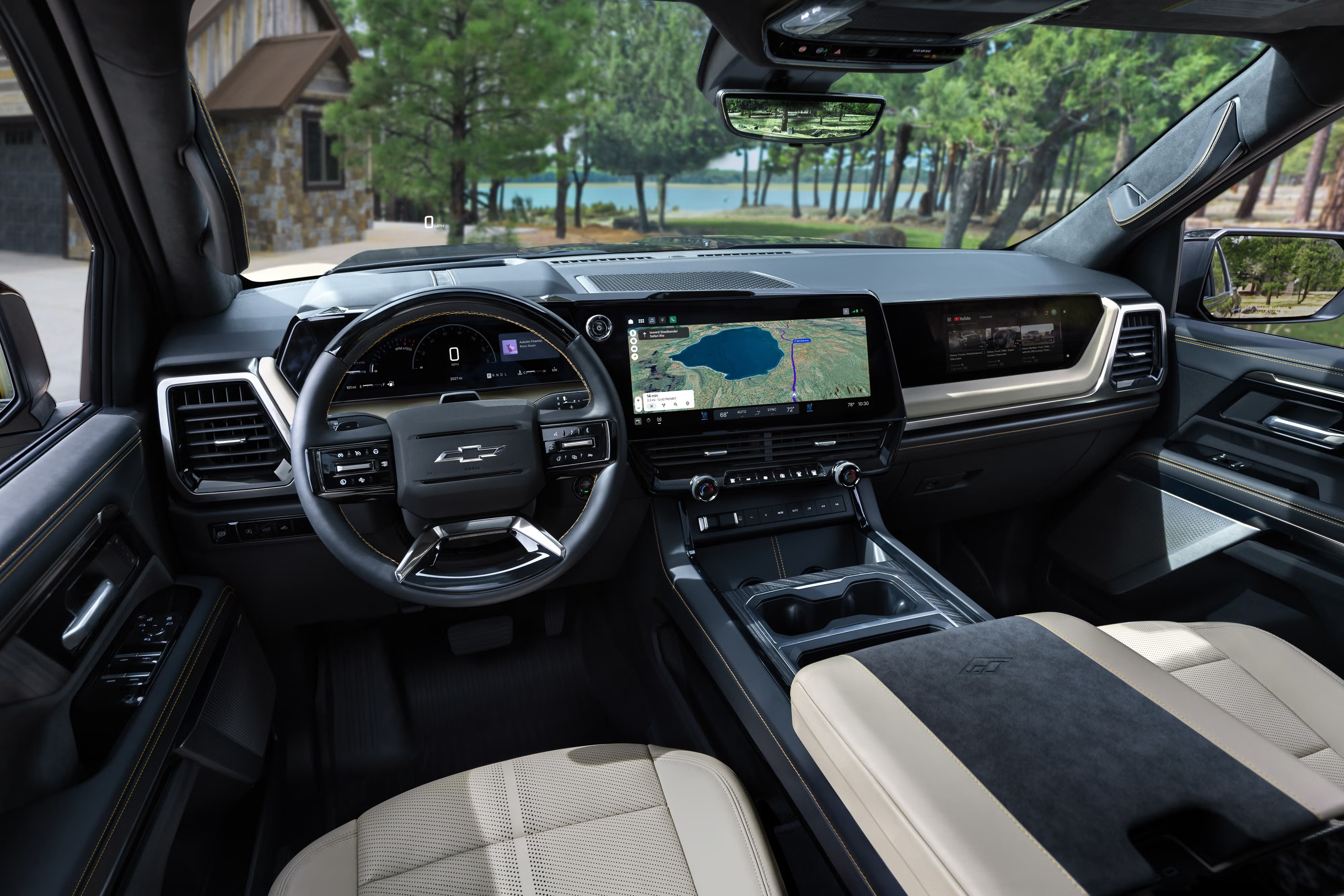 Interior de um carro moderno com volante preto e logotipo Chevrolet, painel digital e tela central exibindo um mapa de navega&ccedil;&atilde;o. Bancos bege e preto, console central com porta-copos. Ao fundo, uma casa de madeira e um lago cercado por &aacute;rvores.