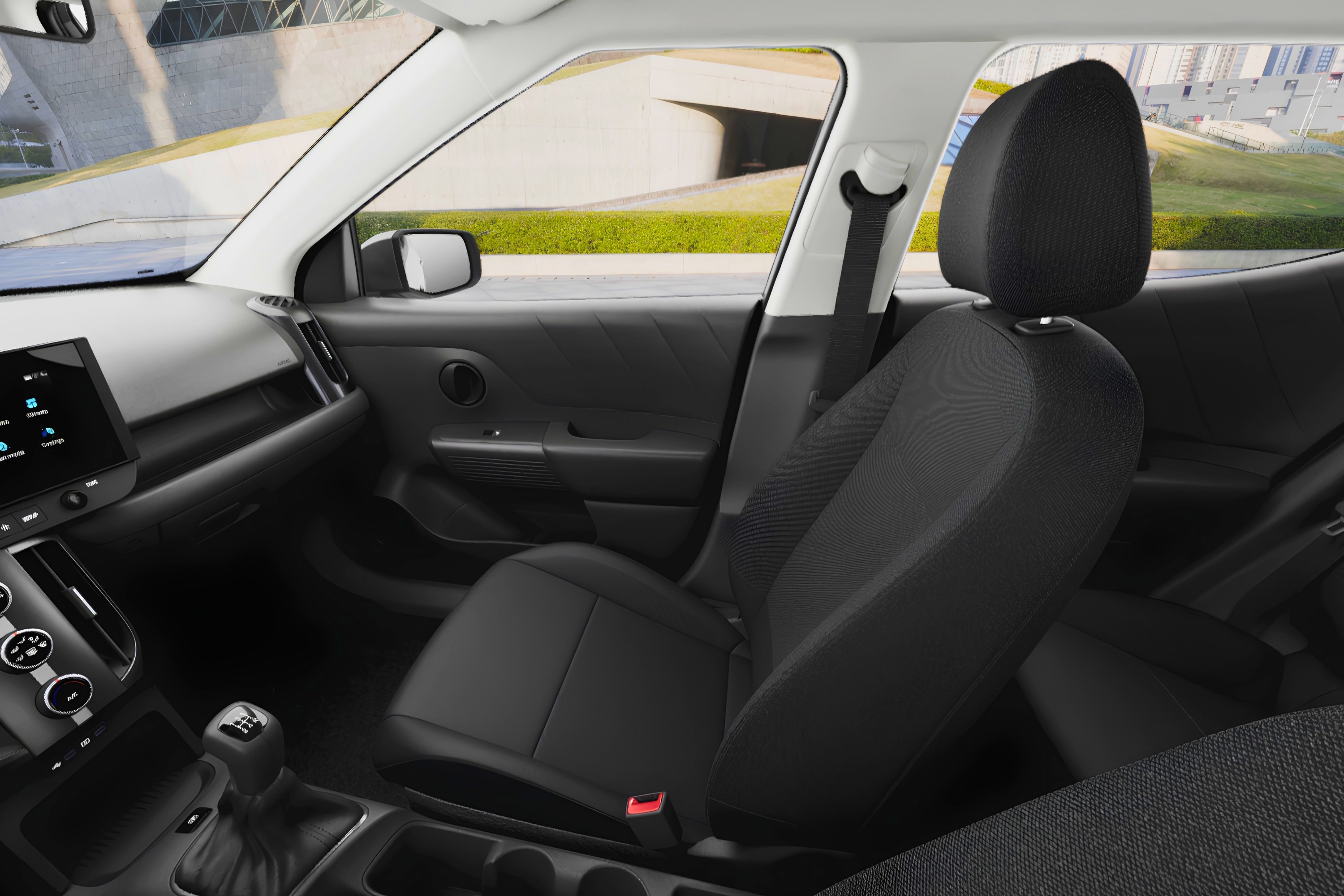 Interior de um carro, mostrando o banco do passageiro dianteiro em tecido preto, o painel com tela multim&iacute;dia e c&acirc;mbio manual. Ao fundo, uma estrada com gramado verde e edif&iacute;cios modernos