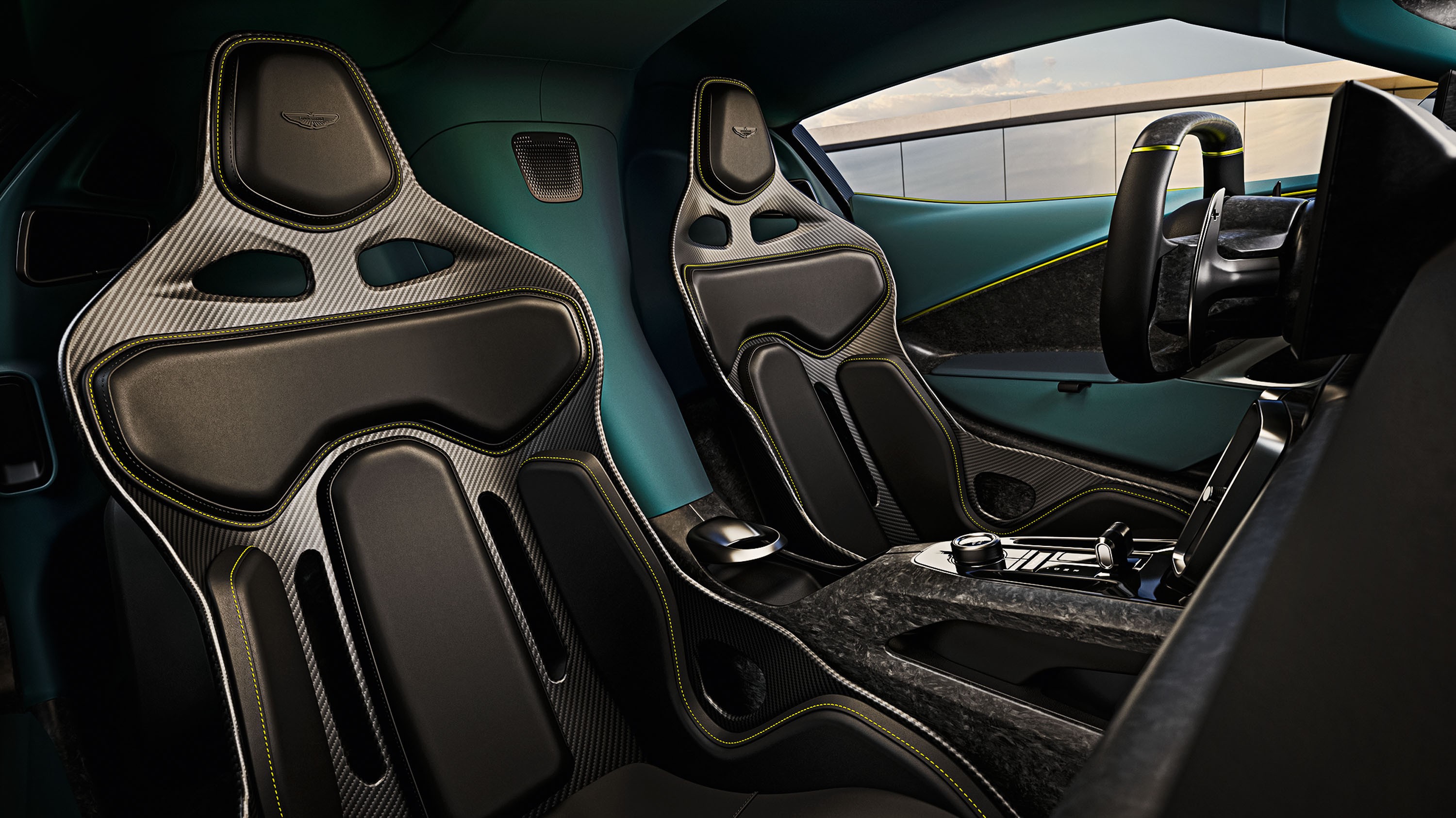 Interior de carro esportivo com dois bancos concha pretos e cinzas de fibra de carbono, costura amarela. Painel e portas em tom verde-azulado, volante &agrave; direita. Console central com detalhes em fibra de carbono e bot&otilde;es prateados. Janela lateral exibe um c&eacute;u claro e uma parede cinza ao fundo