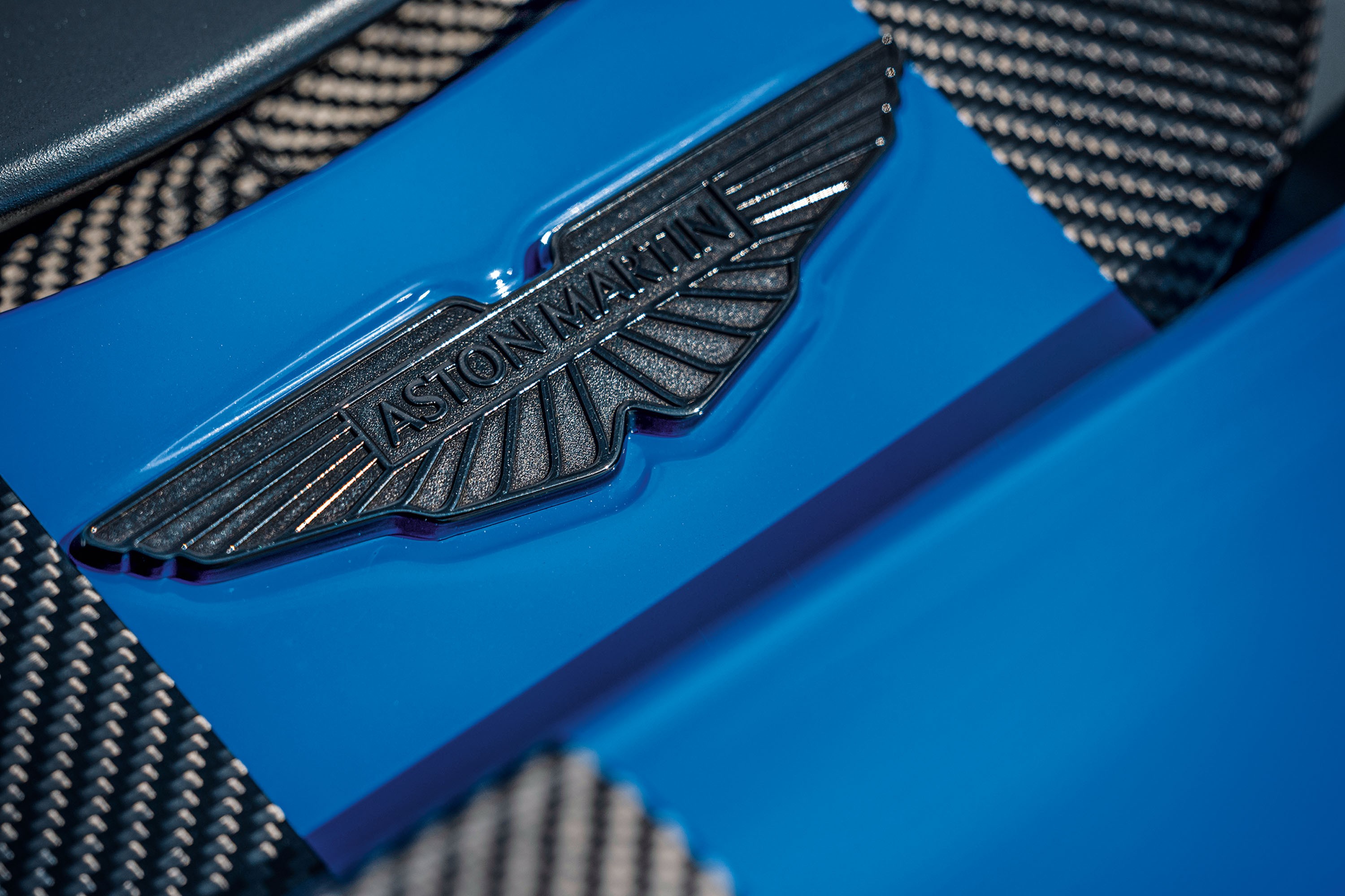 Emblema met&aacute;lico cinza escuro da Aston Martin com asas e o nome da marca em relevo, sobre uma superf&iacute;cie azul brilhante e um tecido quadriculado cinza e branco ao fundo