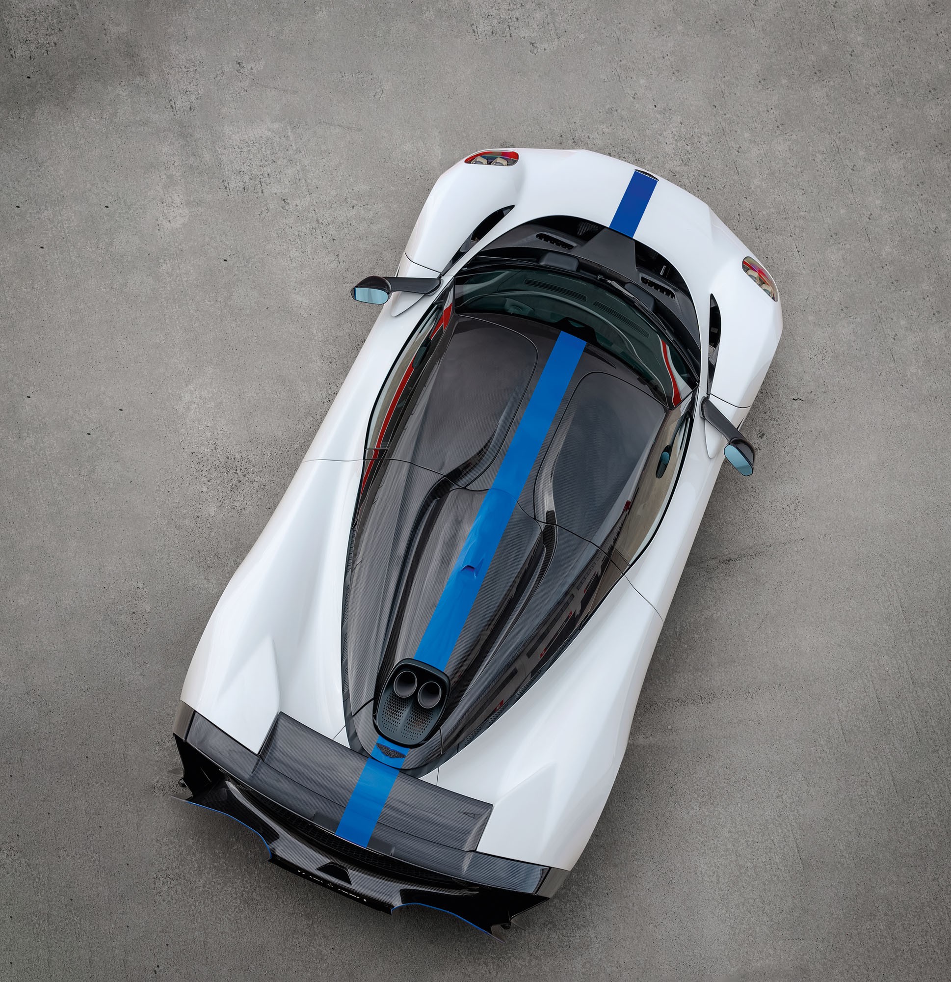Carro esportivo branco com faixa azul central, visto de cima, estacionado em piso de concreto cinza. O teto &eacute; de vidro escuro, e a traseira possui duas sa&iacute;das de escapamento redondas
