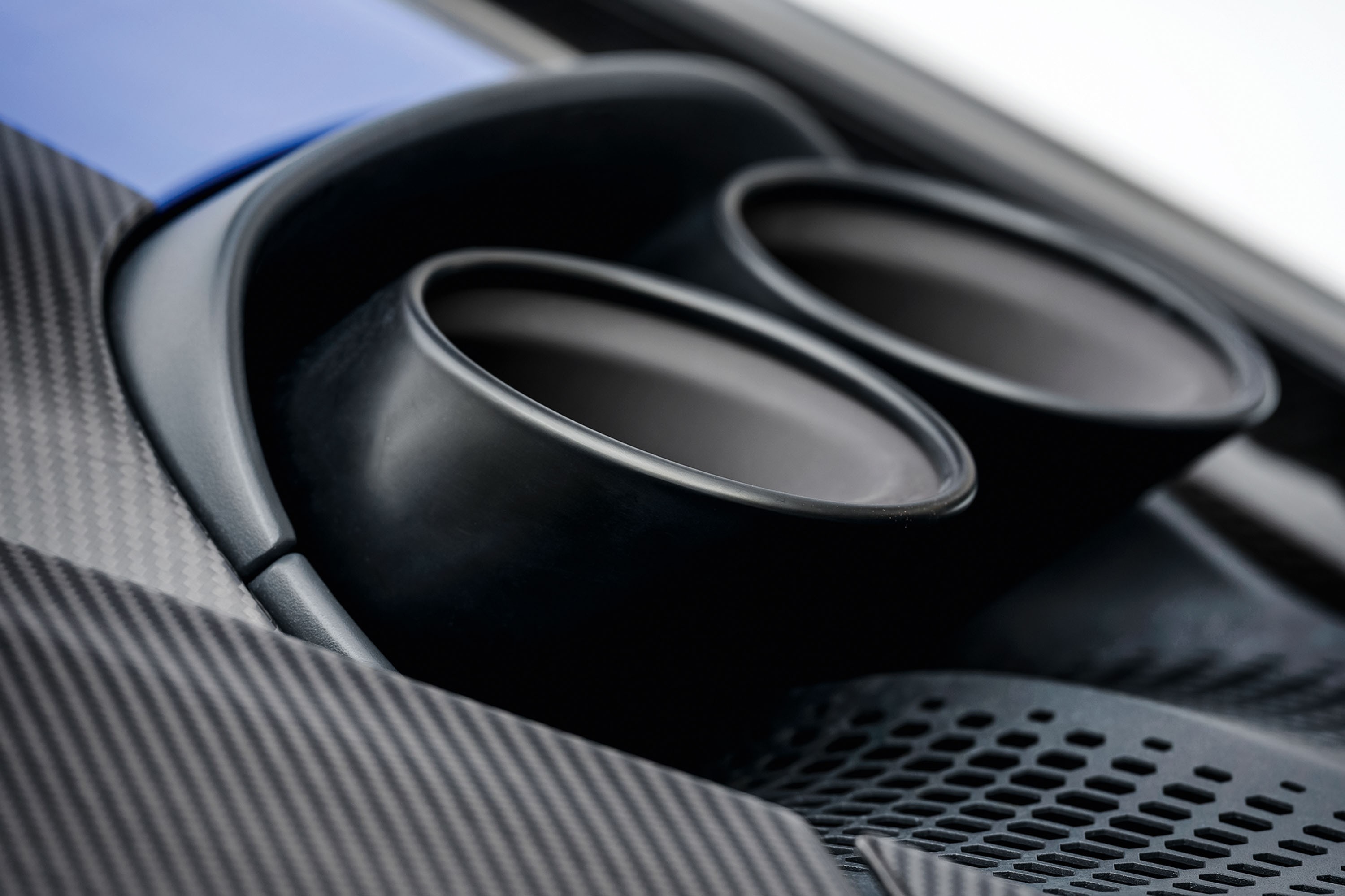Detalhe da traseira de um carro esportivo azul, mostrando duas sa&iacute;das de escapamento pretas foscas e parte da carroceria em fibra de carbono com textura diagonal. Abaixo dos escapamentos, uma grade preta com padr&atilde;o hexagonal.