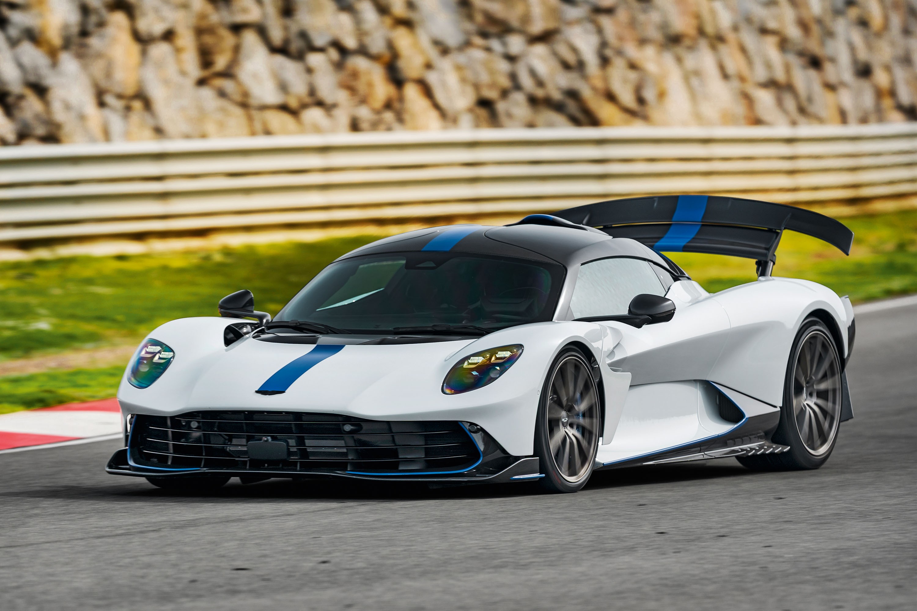 Carro esportivo branco com teto e detalhes azuis e pretos, em uma pista de corrida com barreiras brancas e grama ao fundo
