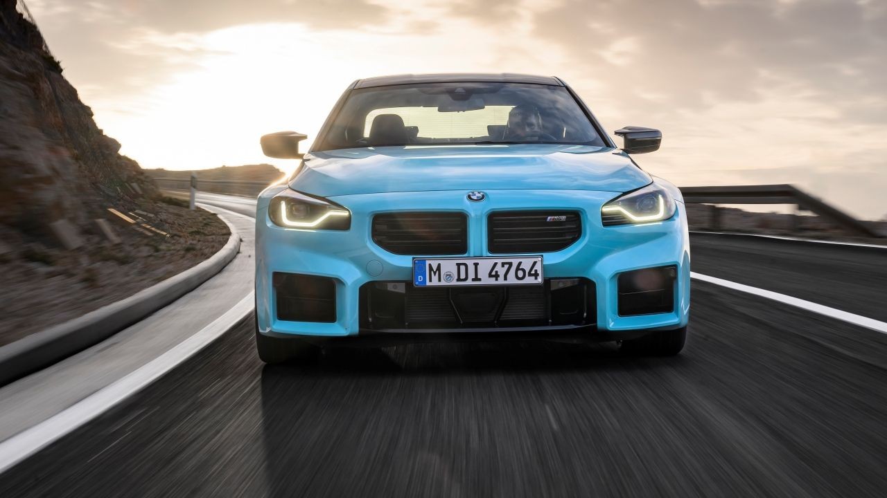 Carro esportivo BMW M2 azul claro em uma estrada sinuosa, visto de frente, com far&oacute;is acesos e p&ocirc;r do sol ao fundo