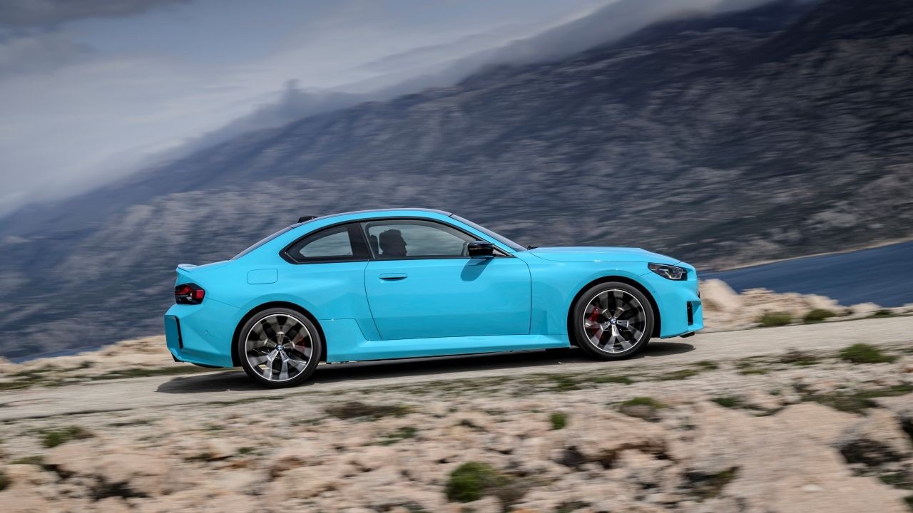 Carro esportivo azul-claro, modelo cup&ecirc;, em movimento numa estrada de terra, com montanhas escuras e um lago azul ao fundo