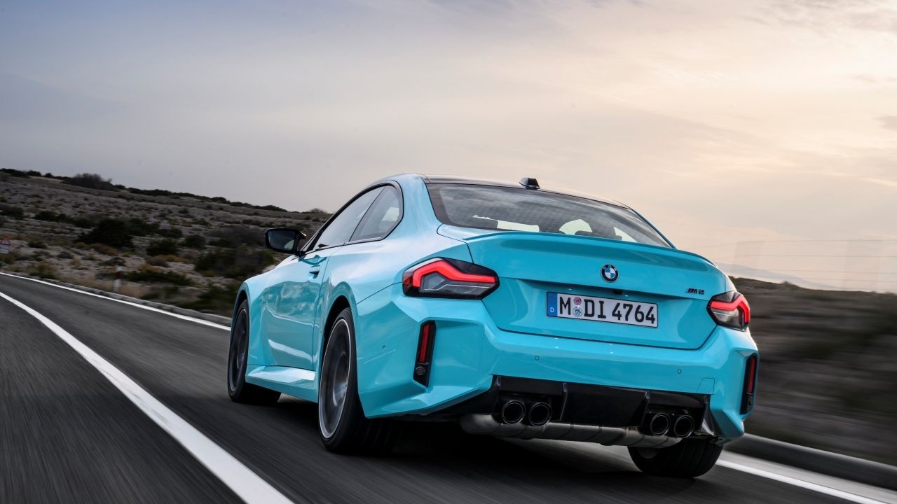 Carro esportivo BMW M2 azul-turquesa visto por tr&aacute;s, acelerando em uma estrada sinuosa com montanhas &aacute;ridas ao fundo e c&eacute;u nublado ao entardecer