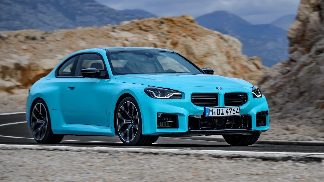 Carro esportivo BMW M2 azul-turquesa, com teto preto, parado em uma estrada de asfalto. Ao fundo, montanhas rochosas e c&eacute;u azul claro. A placa do carro &eacute; M DI 4764