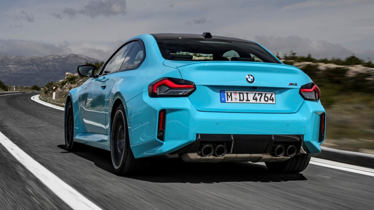 Carro esportivo BMW M2 azul-claro, visto por tr&aacute;s, em uma curva de estrada montanhosa. O ve&iacute;culo tem quatro sa&iacute;das de escapamento e lanternas traseiras escuras, com montanhas e c&eacute;u nublado ao fundo