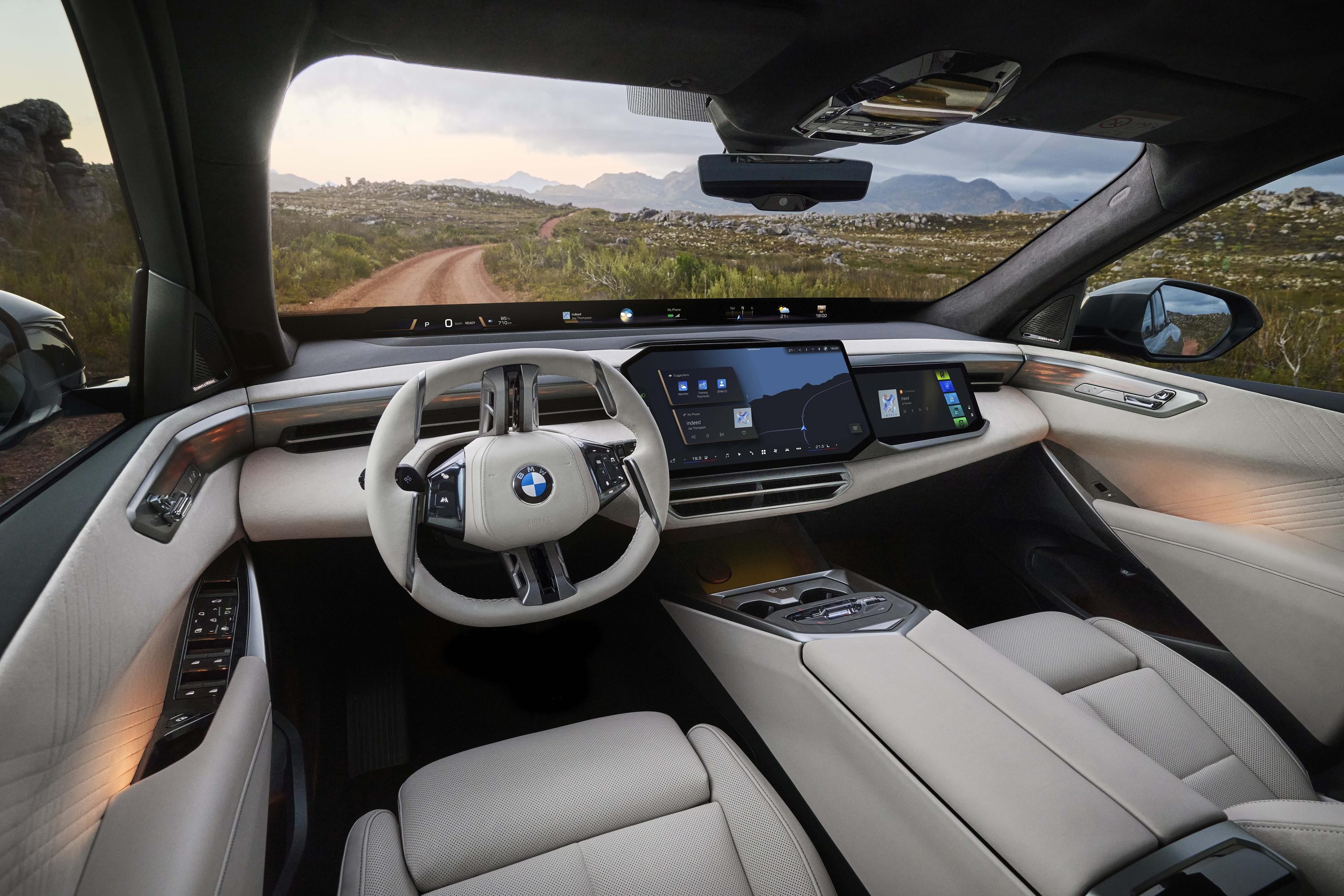 Interior de um carro BMW moderno, com volante branco e o painel digital exibindo um mapa. Ao fundo, uma estrada de terra serpenteia por uma paisagem montanhosa e verdejante sob um c&eacute;u parcialmente nublado