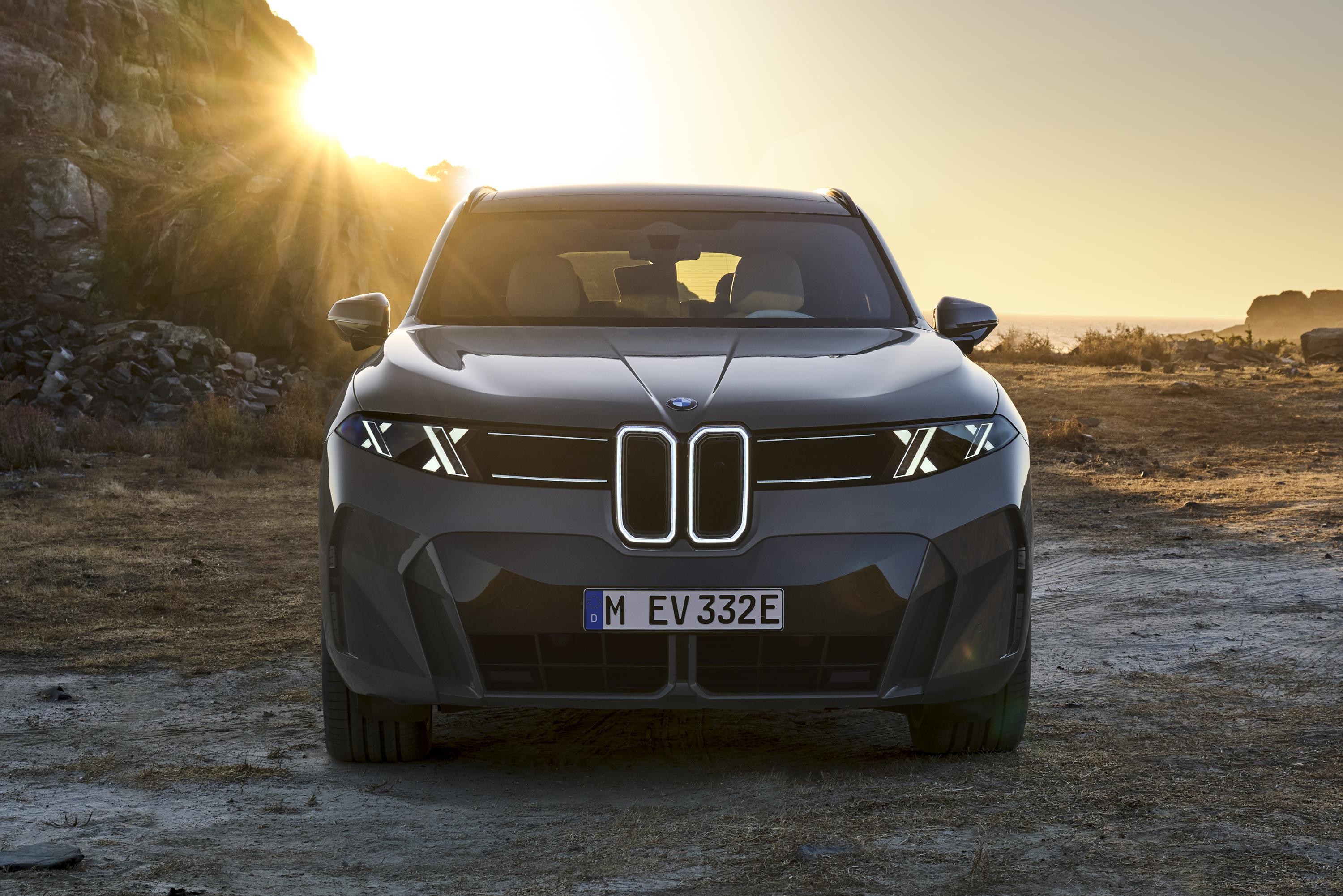 Carro BMW el&eacute;trico cinza escuro, visto de frente, com far&oacute;is em X e grade iluminada, estacionado em terreno &aacute;rido ao p&ocirc;r do sol