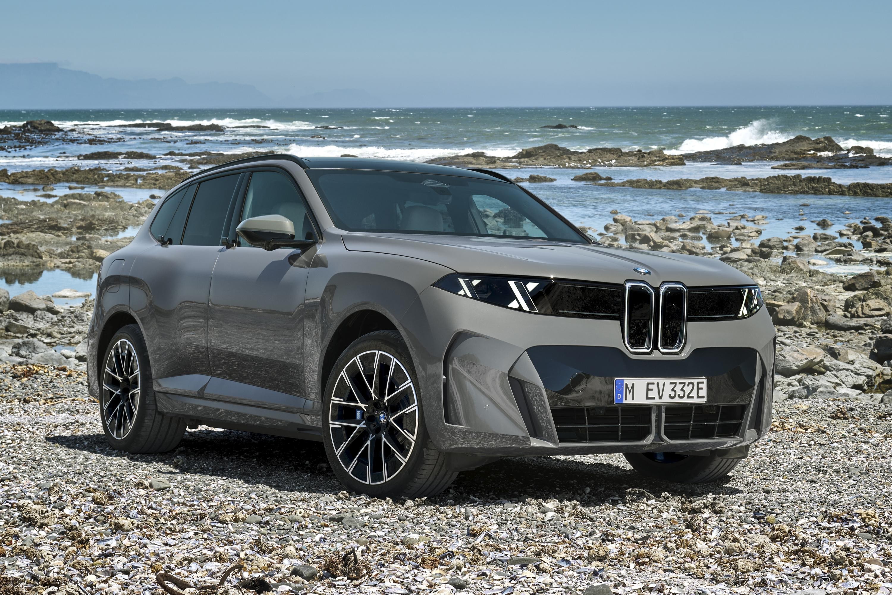 SUV BMW cinza-chumbo estacionado em uma praia rochosa, com o mar azul e montanhas ao fundo. O carro tem far&oacute;is em X e grade frontal dupla preta, com a placa M EV 332E. O c&eacute;u est&aacute; claro e o sol brilha