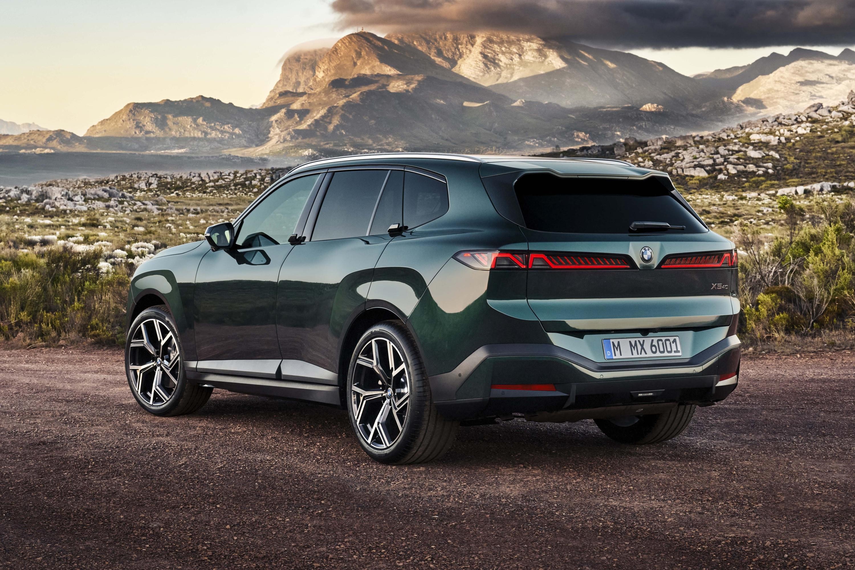 SUV BMW XM verde-escuro, visto de tr&aacute;s e lateral, estacionado em estrada de terra com montanhas e c&eacute;u nublado ao fundo