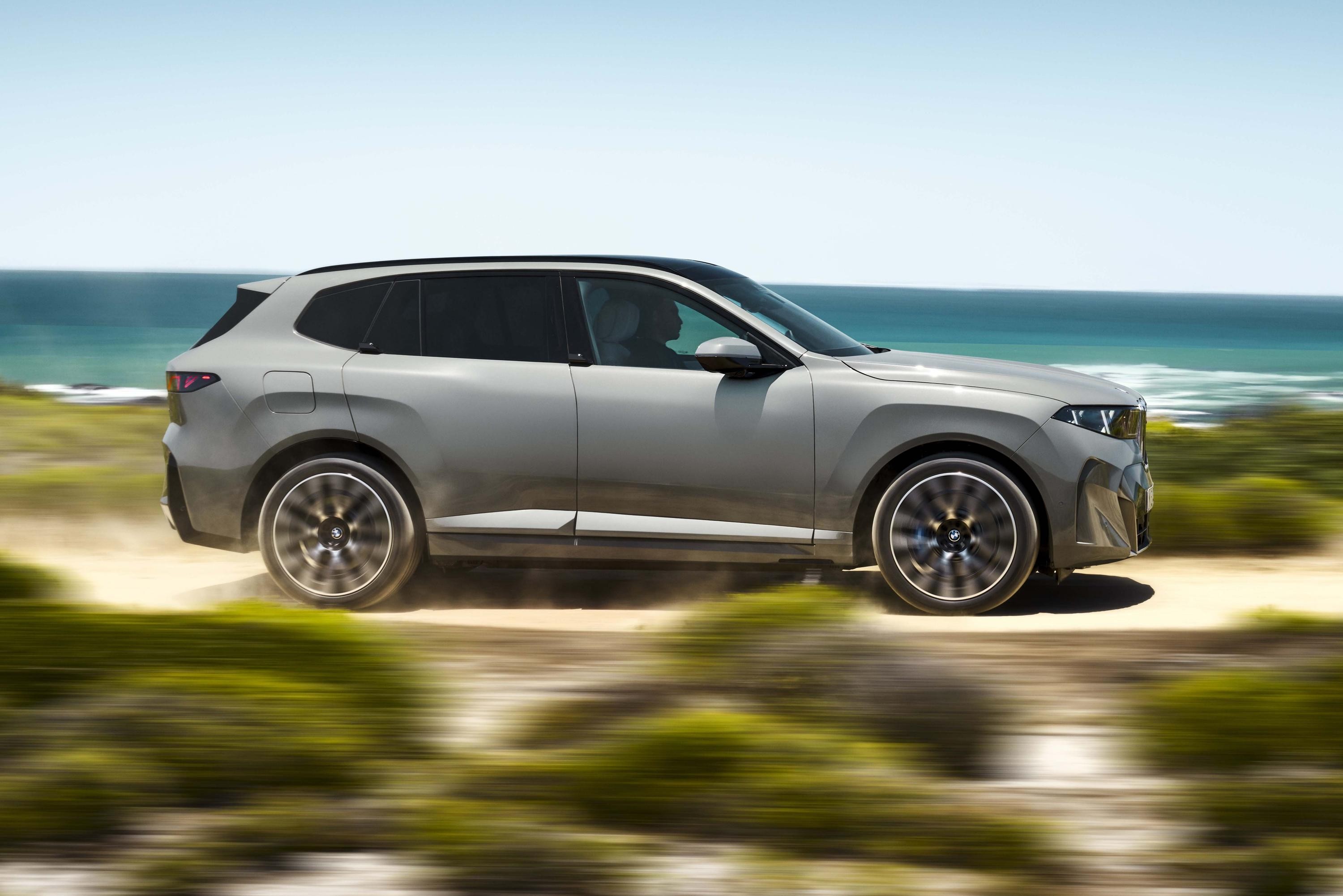 SUV cinza-claro em movimento, visto de lado, com rodas girando rapidamente em uma estrada de terra. Ao fundo, o oceano azul e o c&eacute;u claro, com vegeta&ccedil;&atilde;o borrada em primeiro plano, indicando velocidade