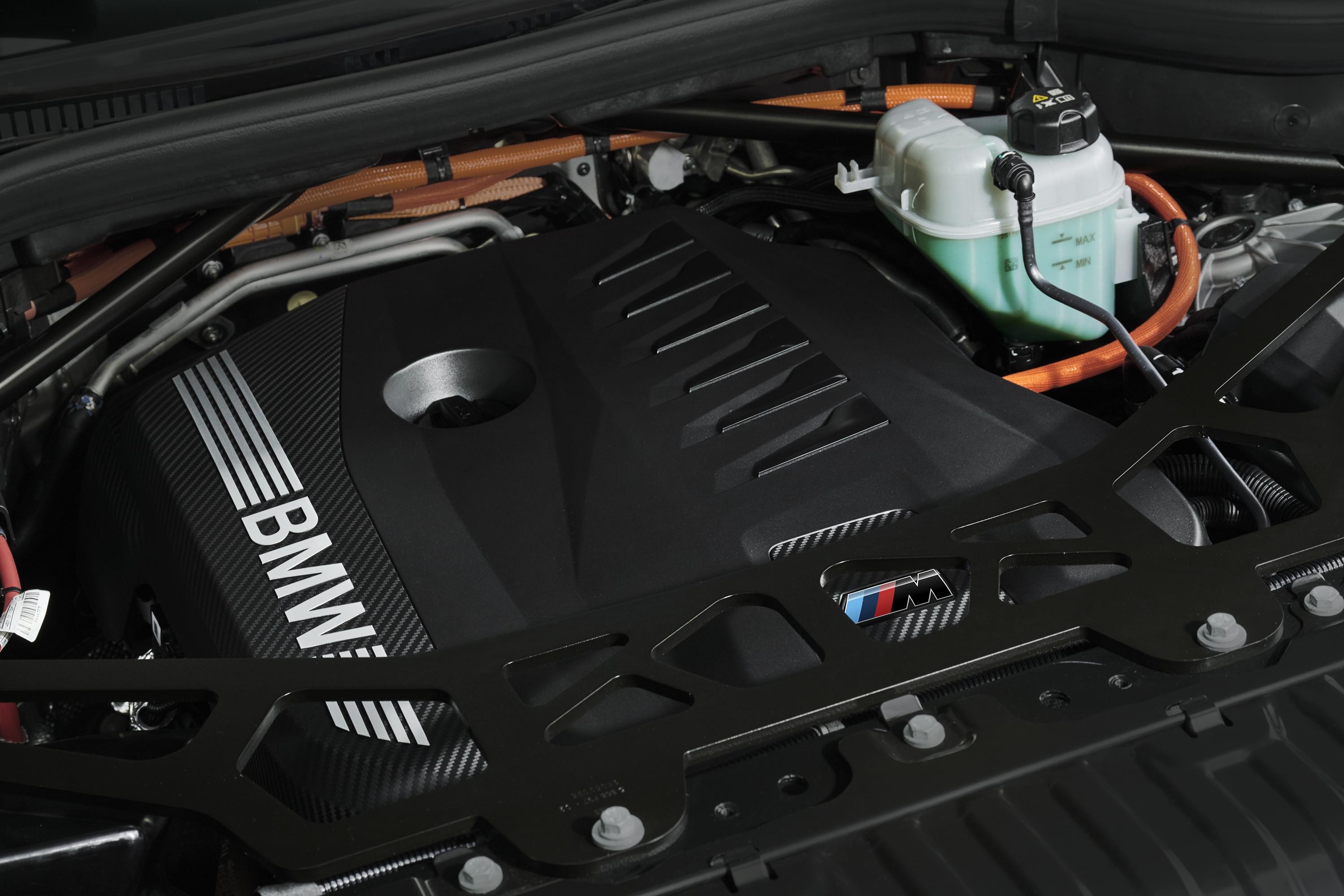 Motor de carro BMW com a tampa preta do motor exibindo o logotipo da marca e um padr&atilde;o de fibra de carbono. Um reservat&oacute;rio de fluido verde claro e tubula&ccedil;&otilde;es laranja s&atilde;o vis&iacute;veis, com uma barra de suporte preta em primeiro plano, que possui o emblema colorido da s&eacute;rie M