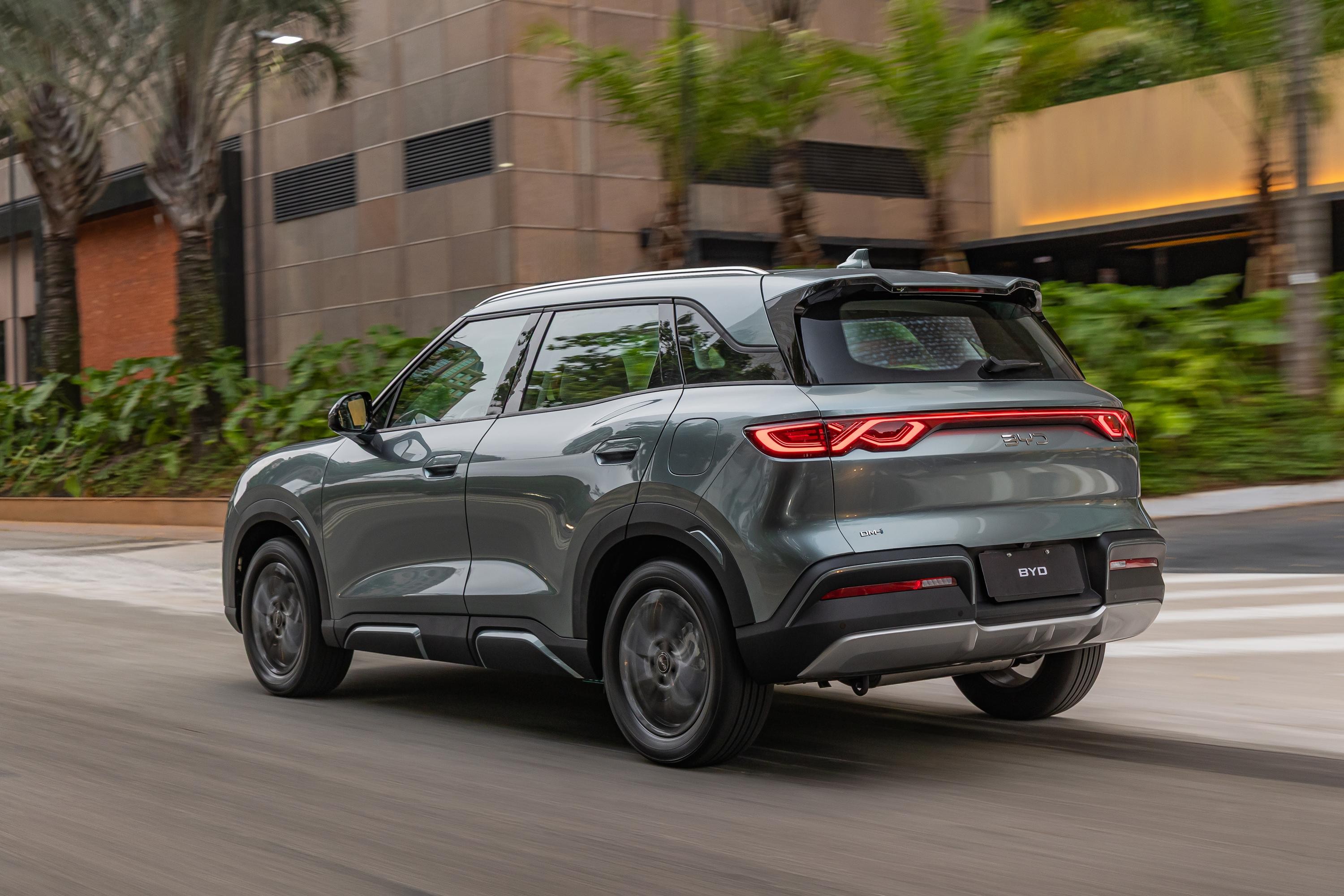 SUV BYD Song Plus DM-i cinza escuro em movimento, visto de tr&aacute;s e lateral, com luzes traseiras acesas. O carro est&aacute; em uma rua asfaltada, com pr&eacute;dios modernos e palmeiras ao fundo, sob luz natural