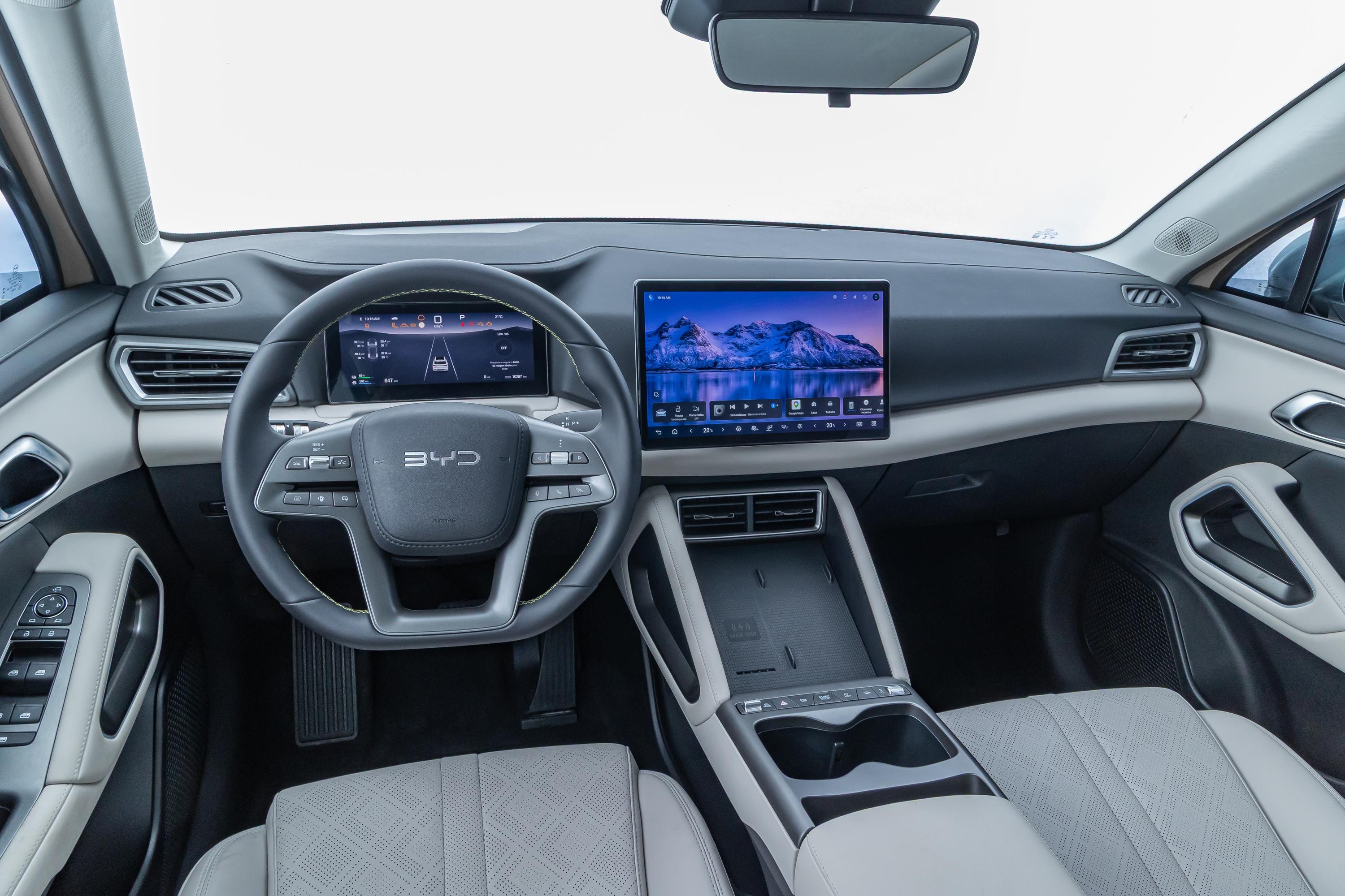 Interior de um carro moderno com volante multifuncional exibindo o logo BYD. O painel possui duas telas digitais: uma menor atr&aacute;s do volante com informa&ccedil;&otilde;es de navega&ccedil;&atilde;o e uma maior central mostrando uma paisagem de montanhas nevadas. Os bancos s&atilde;o cinza claro com detalhes em losango, e o console central tem porta-copos e bot&otilde;es de controle. O acabamento interno combina tons de cinza, preto e branco, com detalhes met&aacute;licos nas ma&ccedil;anetas e sa&iacute;das de ar