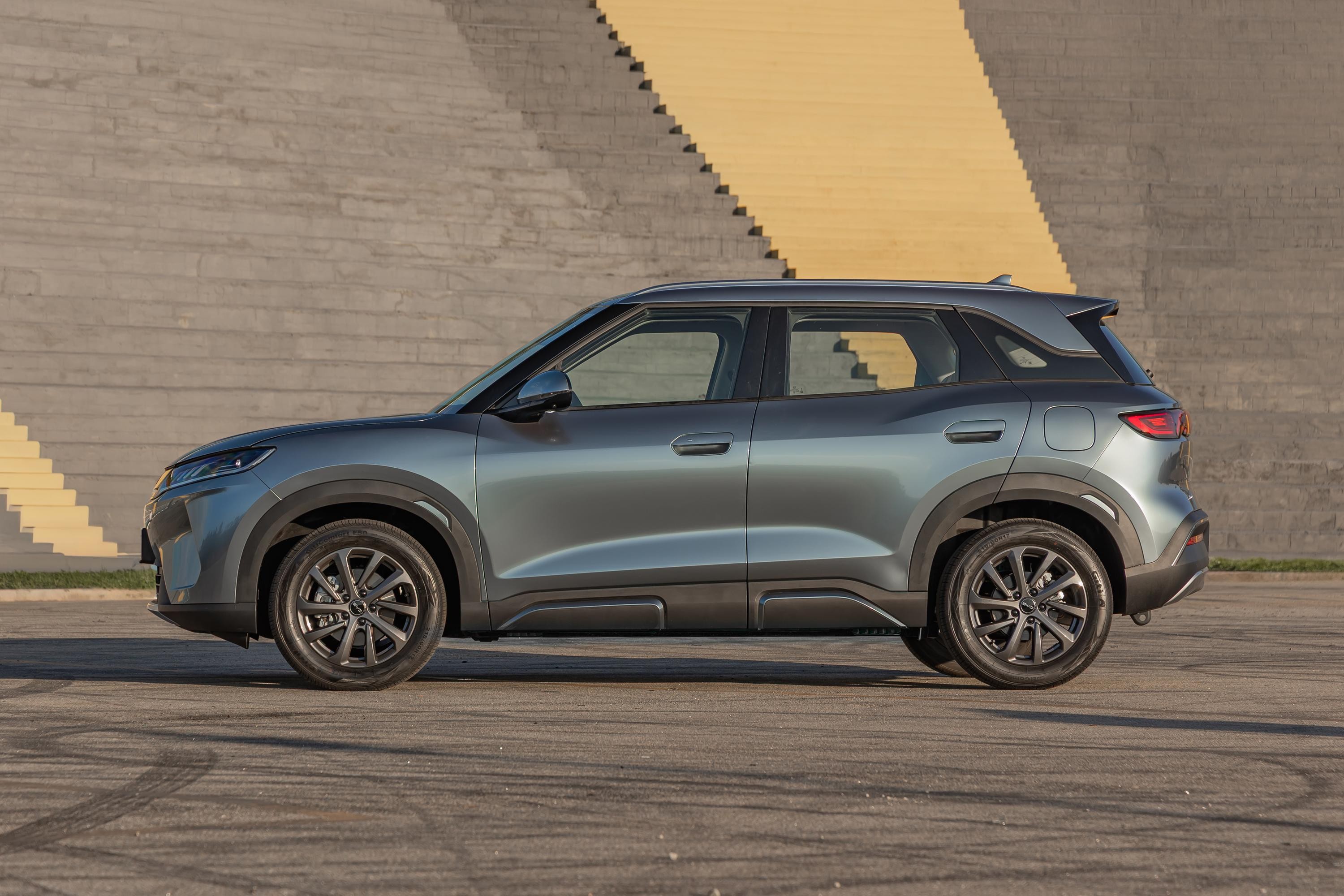 SUV cinza-azulado estacionado de perfil, com rodas escuras e detalhes pretos na parte inferior. Ao fundo, uma parede de concreto com uma escadaria amarela diagonal