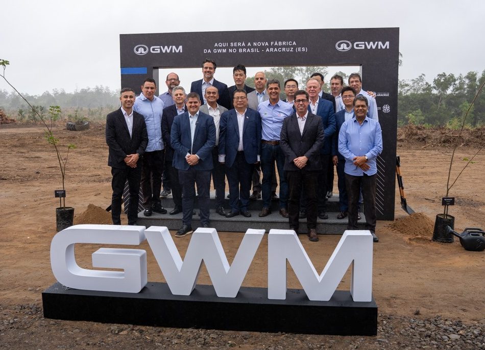Um grupo de cerca de 20 homens, a maioria de terno, posa para foto em um terreno de terra batida. Atr&aacute;s deles, um painel preto com o texto AQUI SER&Aacute; A NOVA F&Aacute;BRICA DA GWM NO BRASIL - ARACRUZ (ES). Em primeiro plano, letras brancas formam GWM