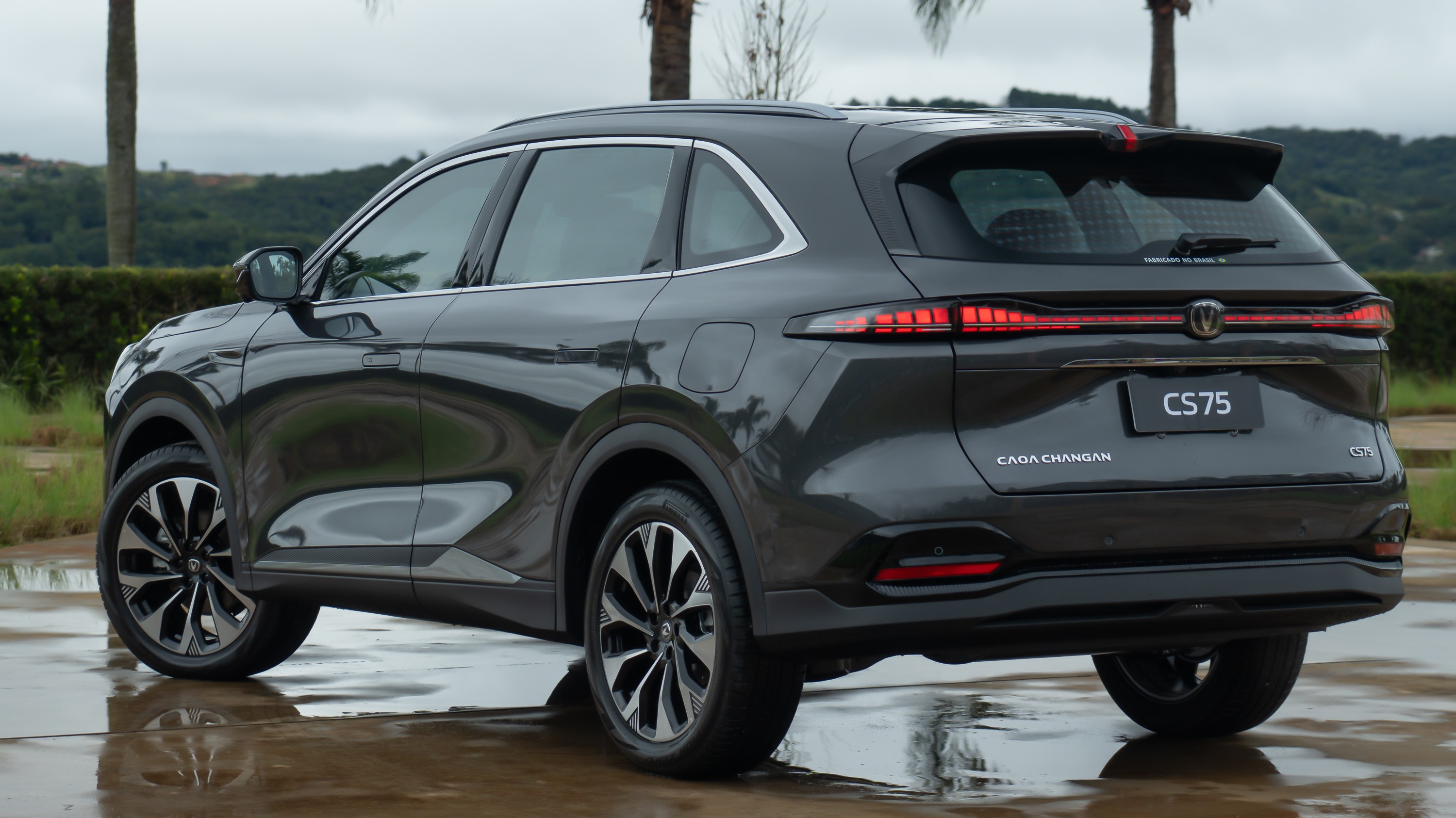 SUV Changan CS75 cinza escuro, visto de traseira e lateral, estacionado em piso molhado com reflexos. Ao fundo, palmeiras e vegeta&ccedil;&atilde;o verde sob c&eacute;u nublado