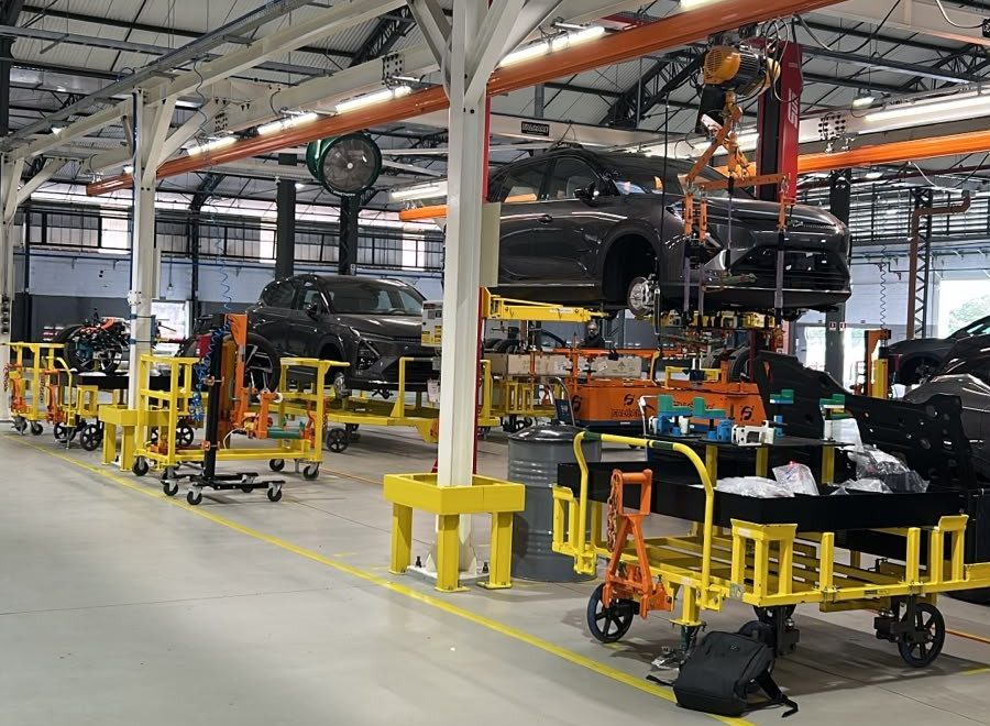 Interior de uma f&aacute;brica de autom&oacute;veis com carros cinzas em diferentes est&aacute;gios de montagem, suspensos por guindastes e em plataformas amarelas. O ch&atilde;o &eacute; cinza claro com faixas amarelas, e o teto alto tem estrutura met&aacute;lica aparente e ilumina&ccedil;&atilde;o industrial. Carrinhos de ferramentas e pe&ccedil;as est&atilde;o espalhados pelo ambiente, indicando um processo de produ&ccedil;&atilde;o ativo