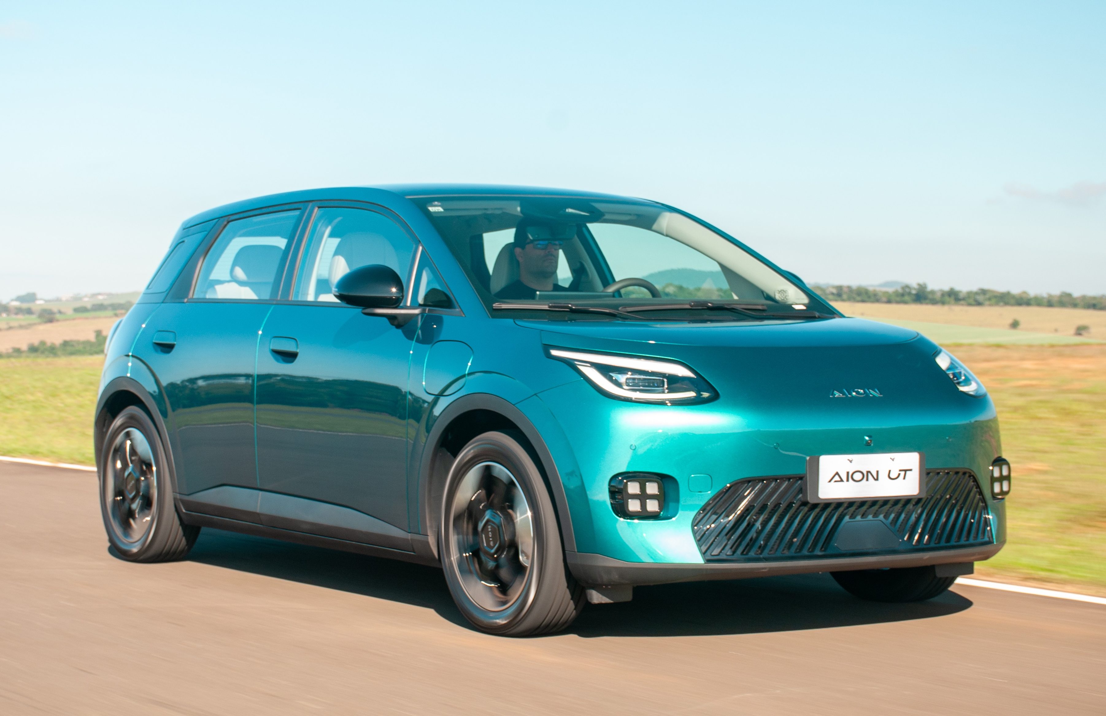 Carro el&eacute;trico Aion UT azul-turquesa em movimento numa estrada asfaltada, com um motorista vis&iacute;vel ao volante. O c&eacute;u azul claro e campos verdes desfocados comp&otilde;em o fundo