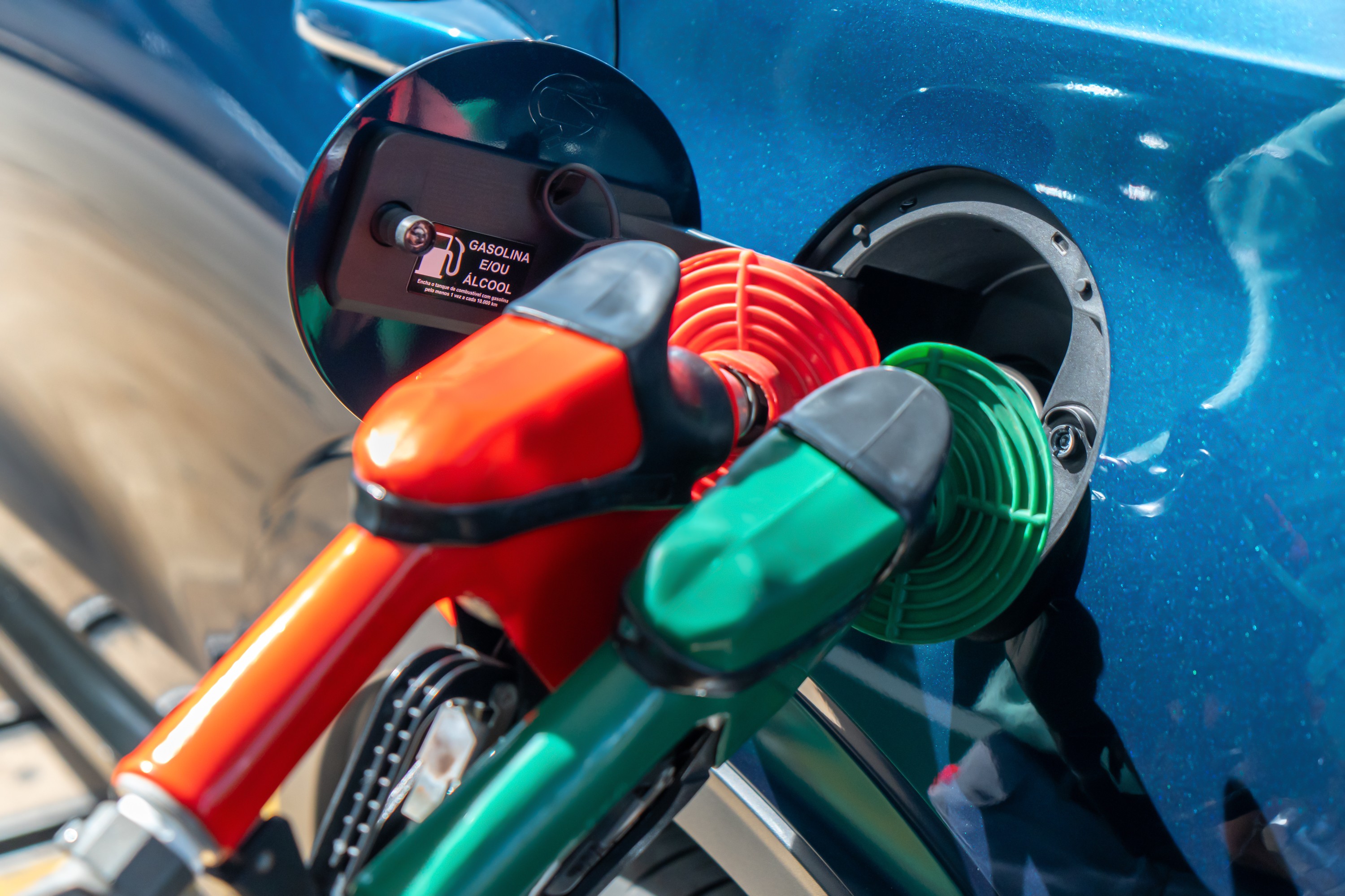 Bicos de bomba de combust&iacute;vel vermelho e verde inseridos no tanque de um carro azul, com a tampa aberta exibindo a inscri&ccedil;&atilde;o GASOLINA E/OU &Aacute;LCOOL