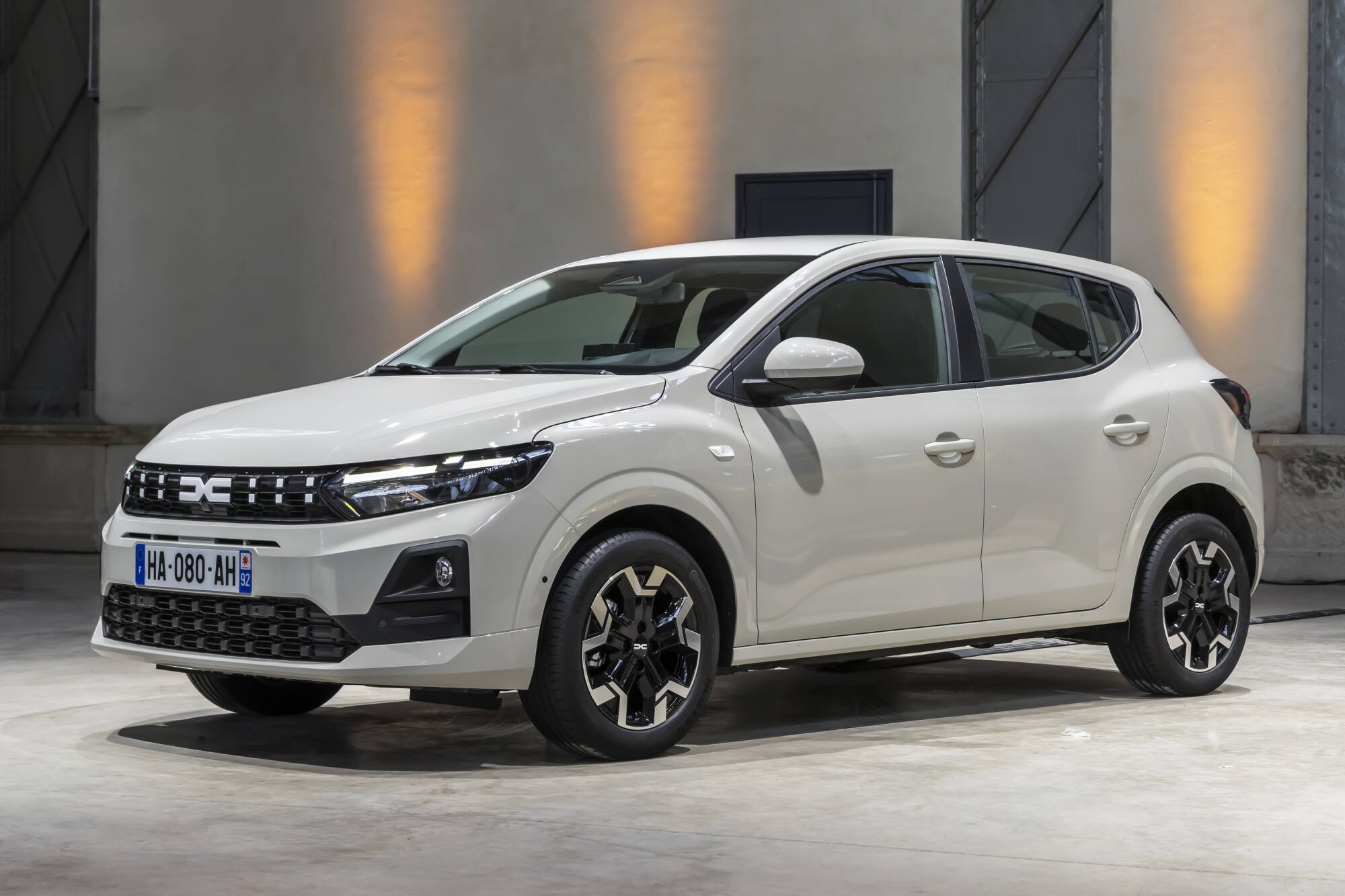 Carro branco Dacia Sandero Stepway, modelo 2024, estacionado em ambiente interno com parede cinza e ilumina&ccedil;&atilde;o amarela, visto de frente e lateral