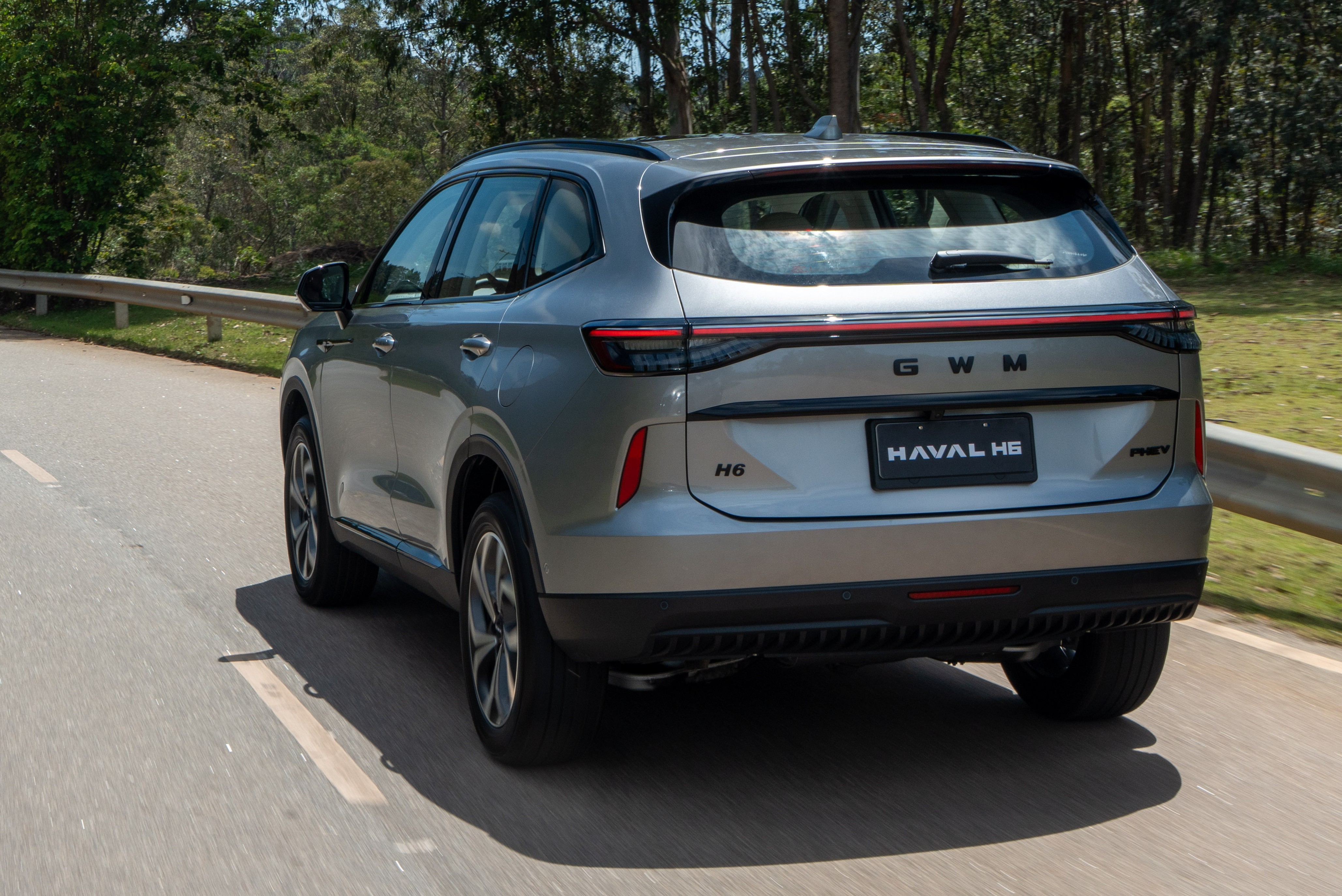 SUV cinza claro, modelo Haval H6, visto por tr&aacute;s, em uma estrada asfaltada com faixa branca. O carro tem lanternas traseiras horizontais e o nome GWM e HAVAL H6 na tampa do porta-malas. H&aacute; &aacute;rvores verdes ao fundo e um guard-rail met&aacute;lico &agrave; direita