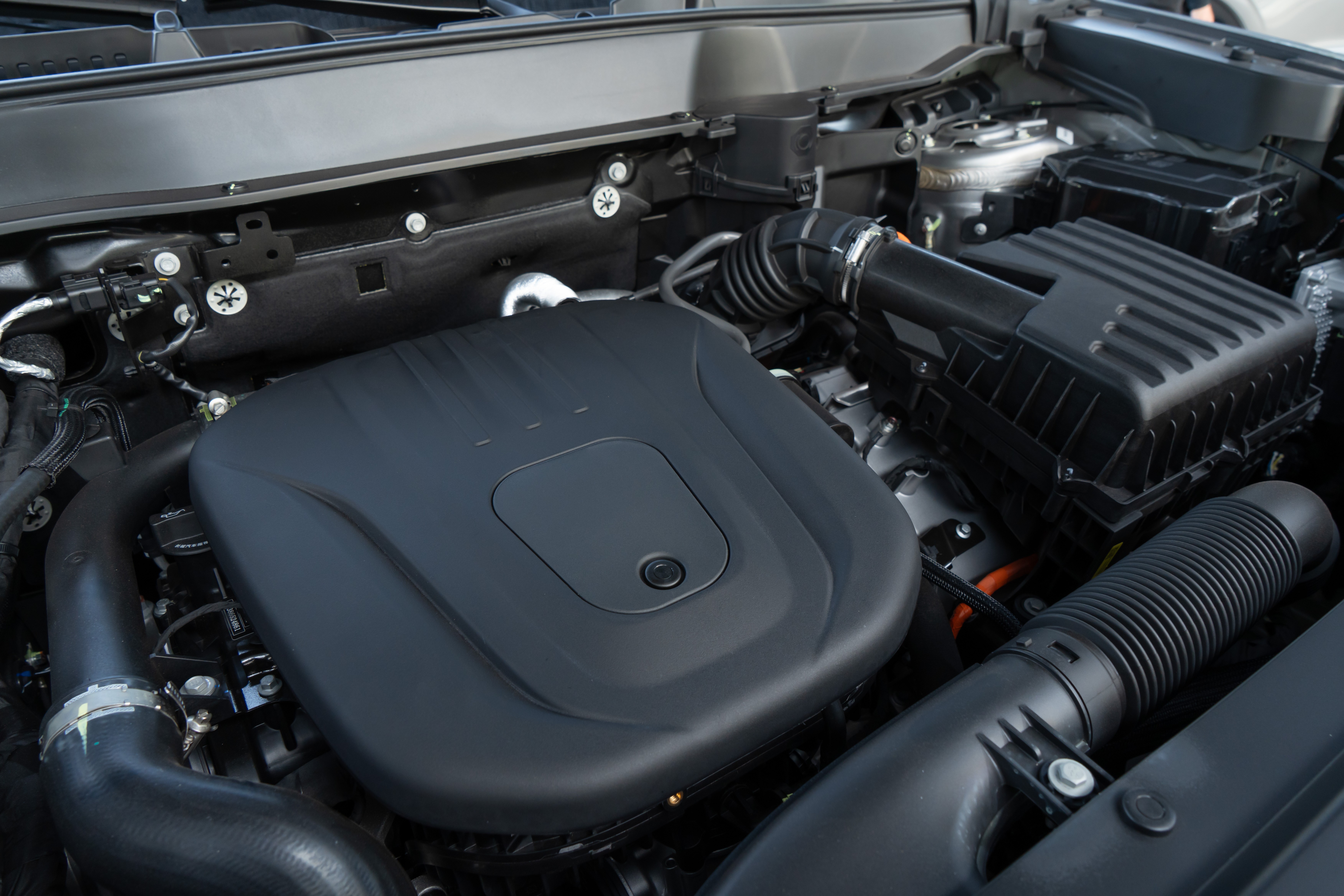 Vista superior do motor de um carro, com a tampa do motor preta e lisa no centro. Ao redor, tubula&ccedil;&otilde;es, fios e componentes mec&acirc;nicos em tons escuros, com detalhes prateados e alaranjados, indicando um ve&iacute;culo moderno