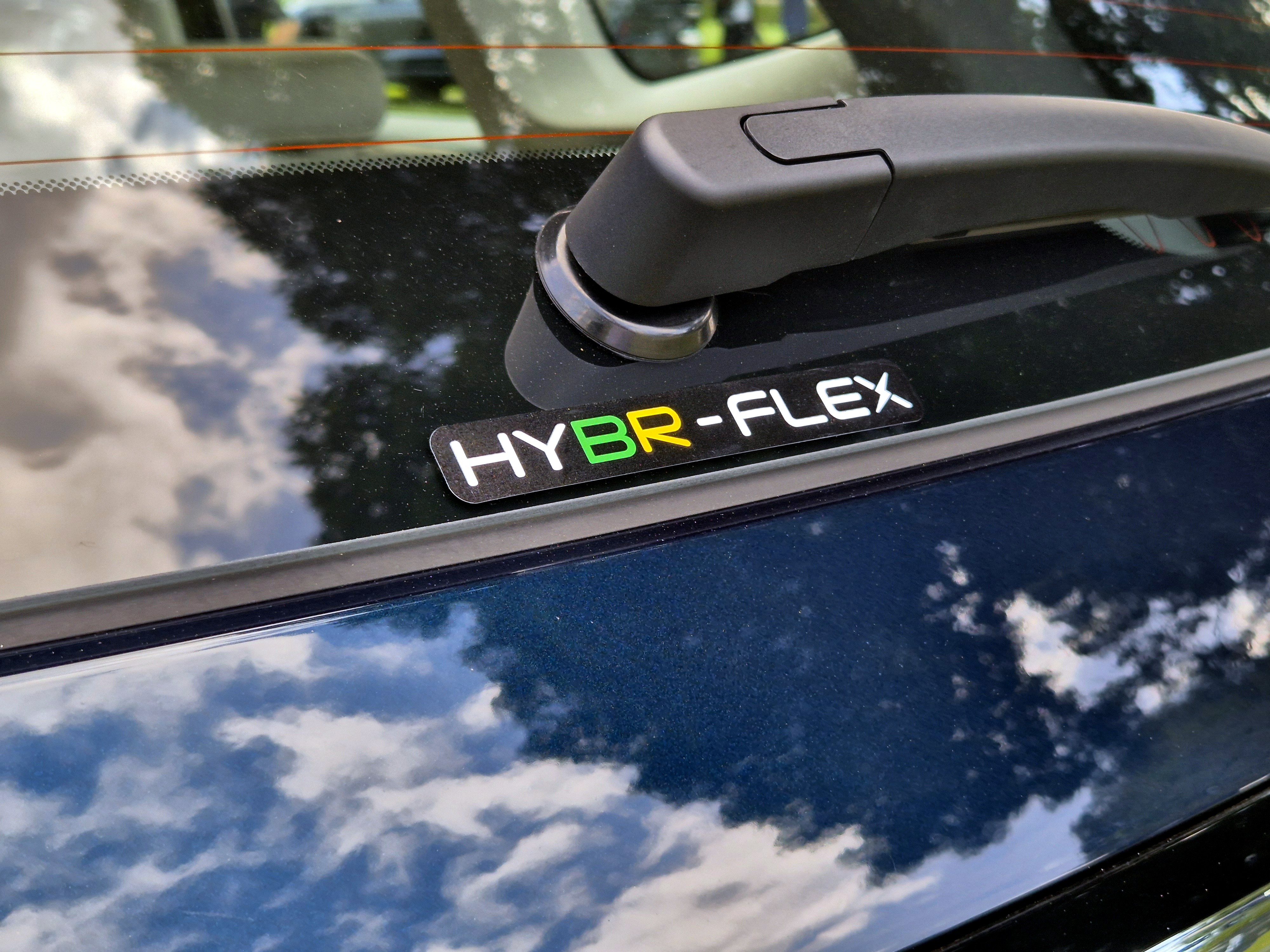 Adesivo HYBR-FLEX em preto com letras brancas, BR em verde e F em amarelo, colado no vidro traseiro de um carro azul escuro, abaixo do limpador de para-brisa. Nuvens e c&eacute;u azul refletem na lataria e no vidro
