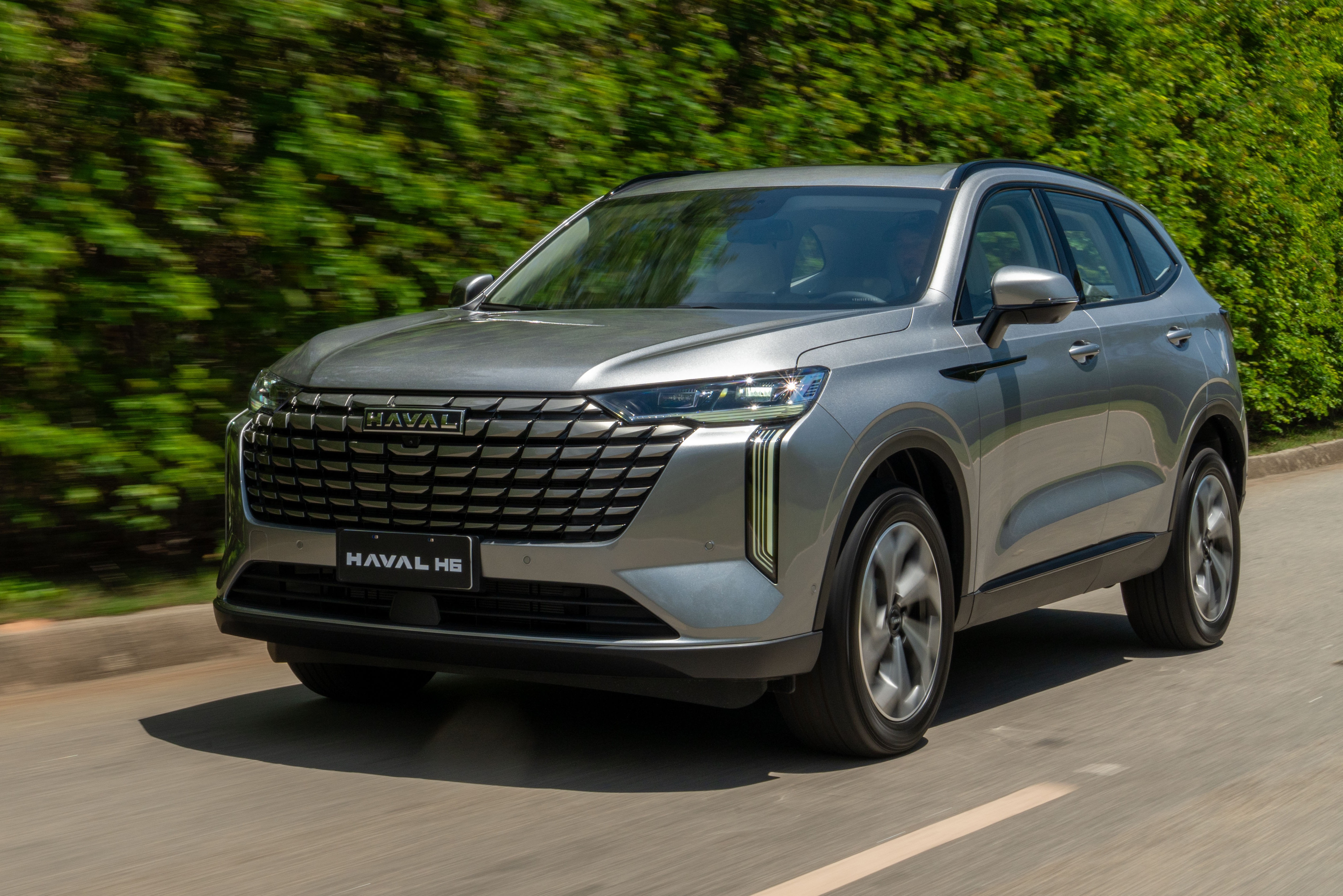SUV Haval H6 cinza-prateado em movimento, visto de frente e levemente de lado, com grade frontal grande e far&oacute;is afilados. O carro est&aacute; em uma estrada asfaltada com faixa branca, e h&aacute; uma vegeta&ccedil;&atilde;o verde borrada ao fundo, indicando velocidade