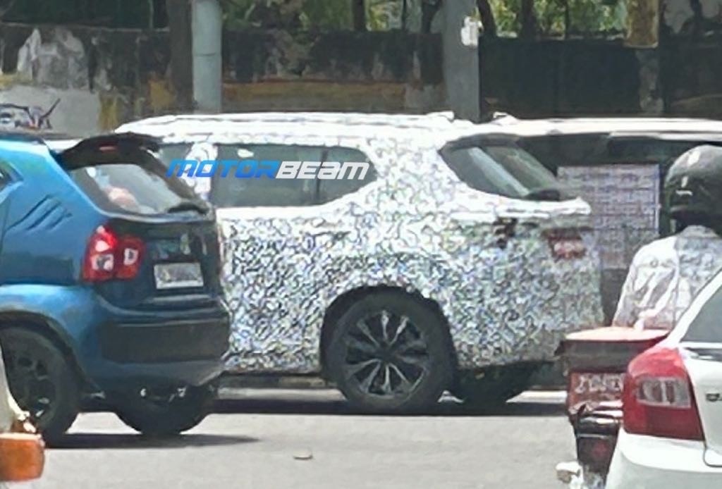 Um SUV camuflado com estampa branca e preta, com rodas escuras, estacionado em uma rua. &Agrave; esquerda, a traseira de um carro azul. &Agrave; direita, a traseira de um carro branco e um motociclista de capacete preto. O fundo &eacute; desfocado com &aacute;rvores e constru&ccedil;&otilde;es