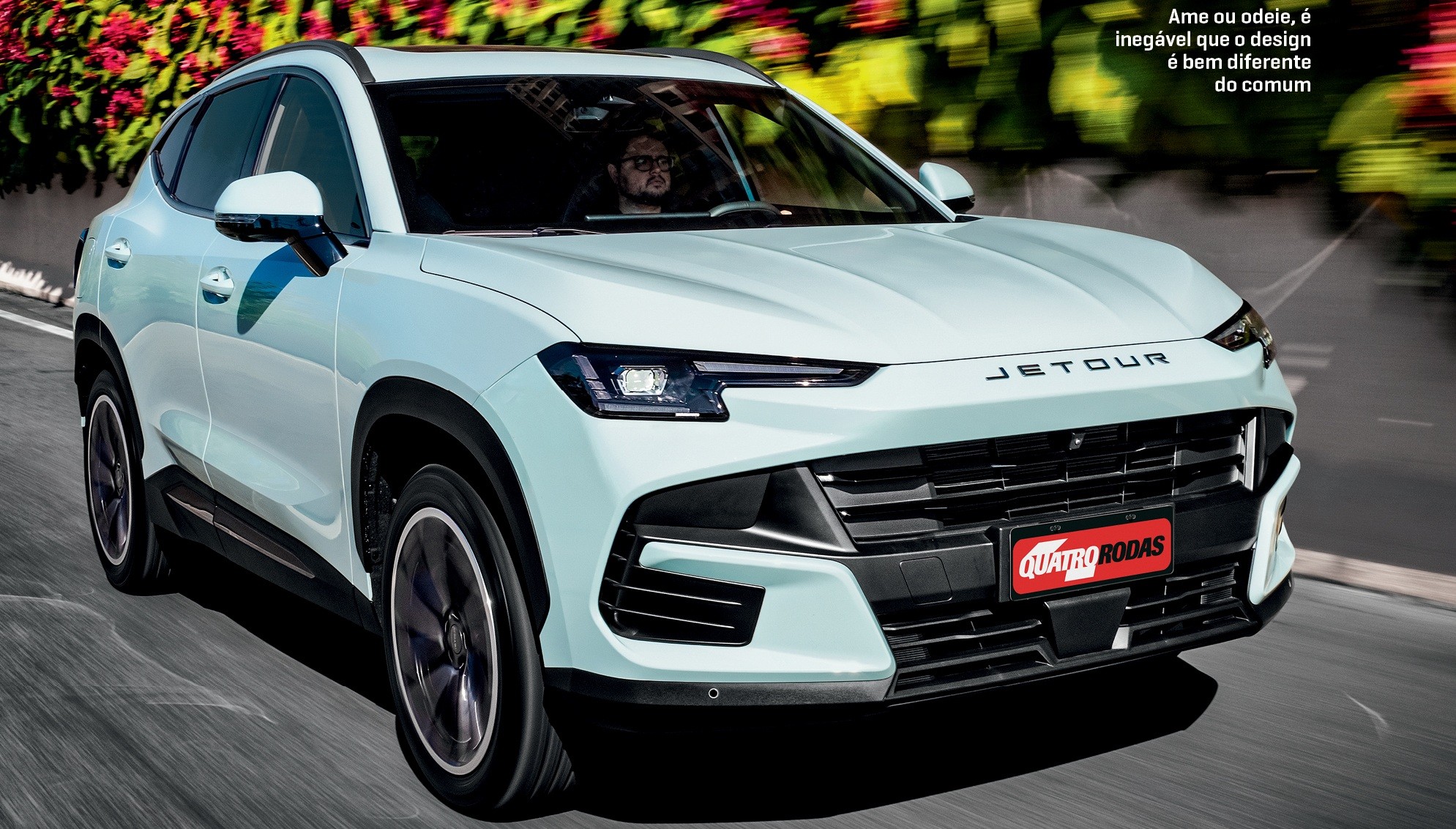 SUV Jetour branco-azulado em movimento na rua, com um homem dirigindo, e vegeta&ccedil;&atilde;o colorida ao fundo. O carro tem o nome JETOUR no cap&ocirc; e um emblema QUATRO RODAS na grade frontal. O texto superior direito diz: Ame ou odeie, &eacute; ineg&aacute;vel que o design &eacute; bem diferente do comum
