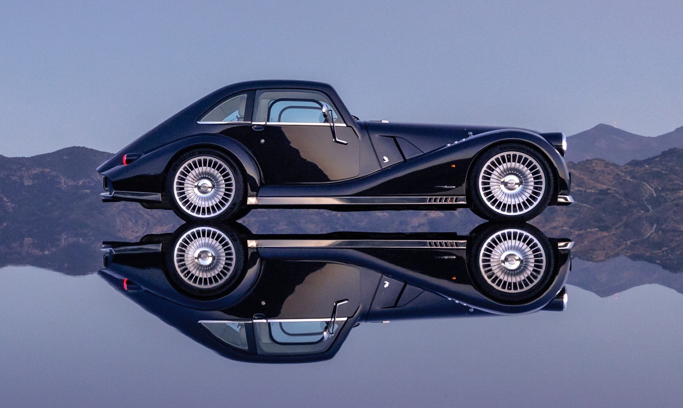 Carro esportivo preto vintage com rodas prateadas, refletido em uma superf&iacute;cie espelhada, com montanhas ao fundo sob c&eacute;u azul claro