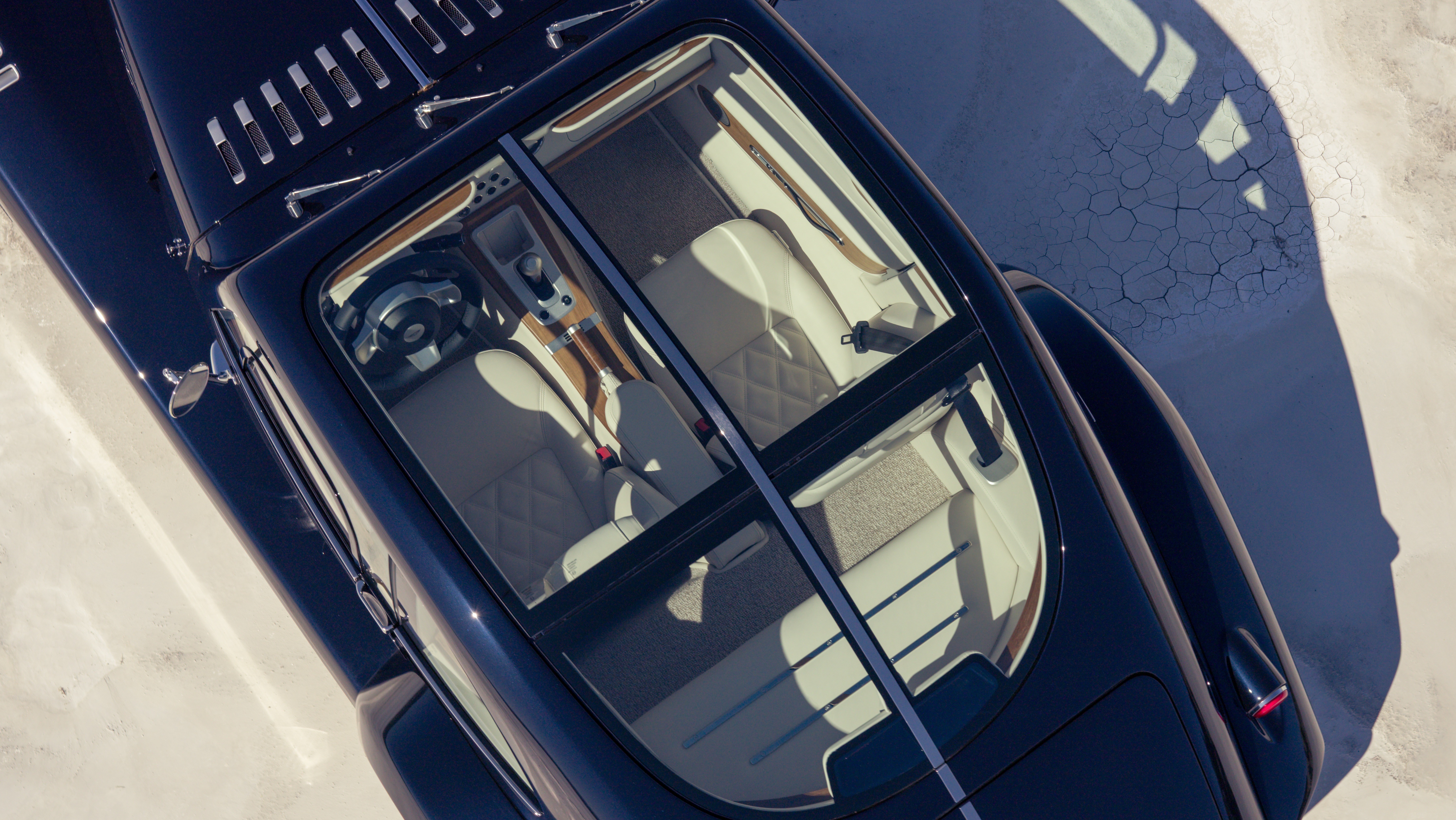 Vista a&eacute;rea de um carro esportivo azul-marinho com teto solar, interior claro e detalhes em madeira, estacionado em uma superf&iacute;cie clara com sombra projetada e rachaduras no ch&atilde;o