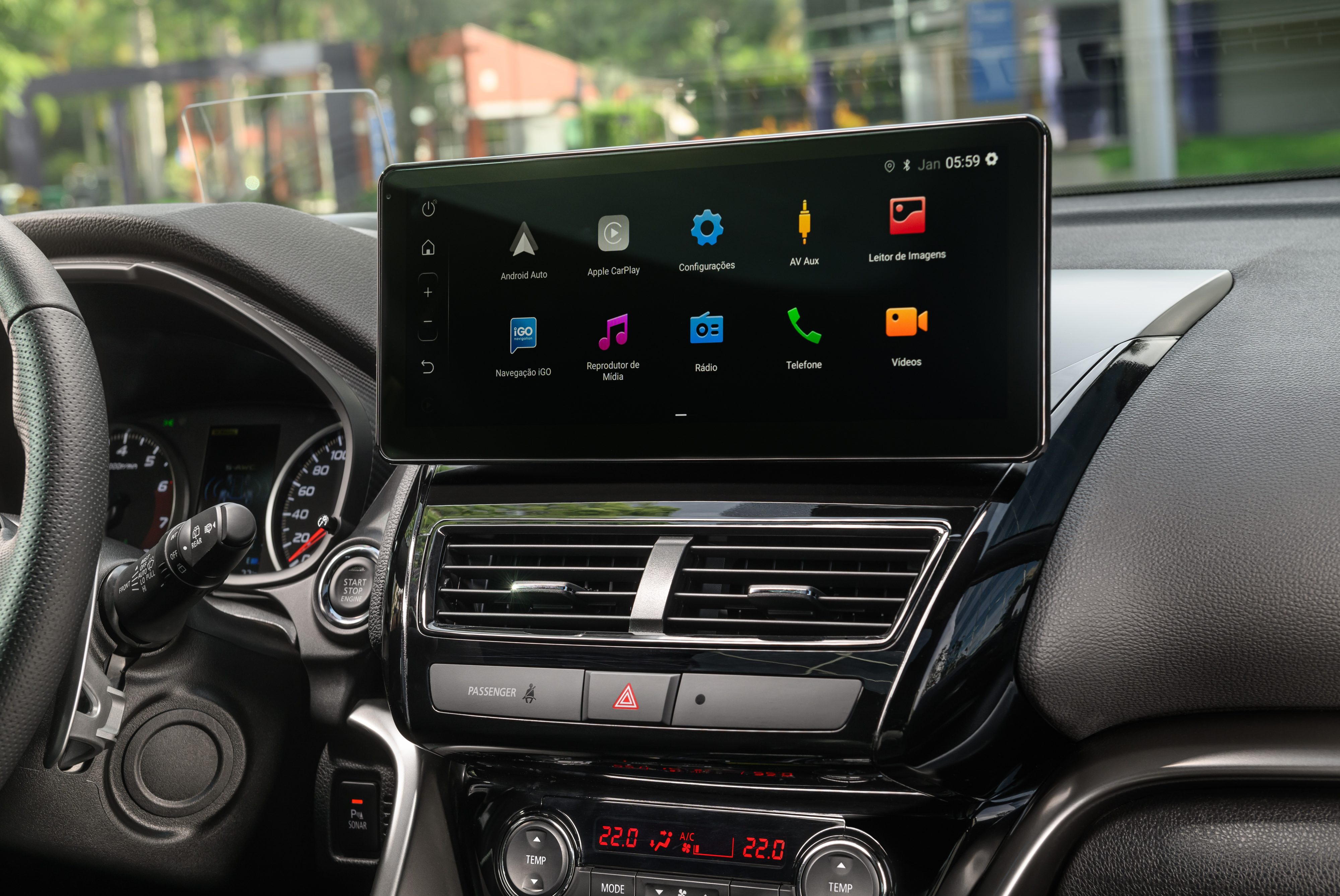 Painel interno de um carro com tela multim&iacute;dia preta exibindo &iacute;cones de aplicativos como Android Auto, Apple CarPlay, Configura&ccedil;&otilde;es, R&aacute;dio e Telefone. Abaixo, sa&iacute;das de ar e controles de temperatura digitais marcando 22.0 graus. Volante e parte do painel de instrumentos s&atilde;o vis&iacute;veis &agrave; esquerda