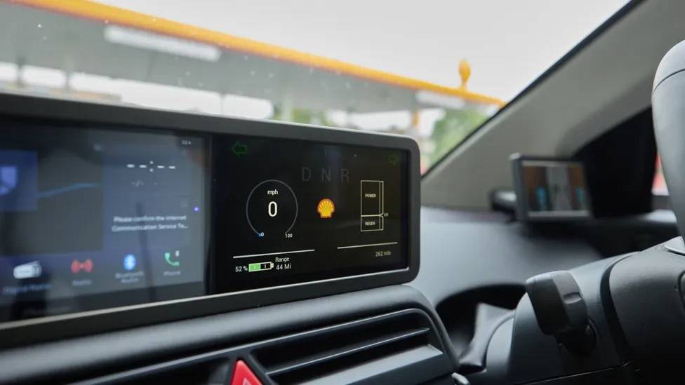 Painel digital de carro com veloc&iacute;metro marcando zero, logo da Shell, indicador de bateria em 52% e autonomia de 44 milhas. Ao fundo, um posto de gasolina