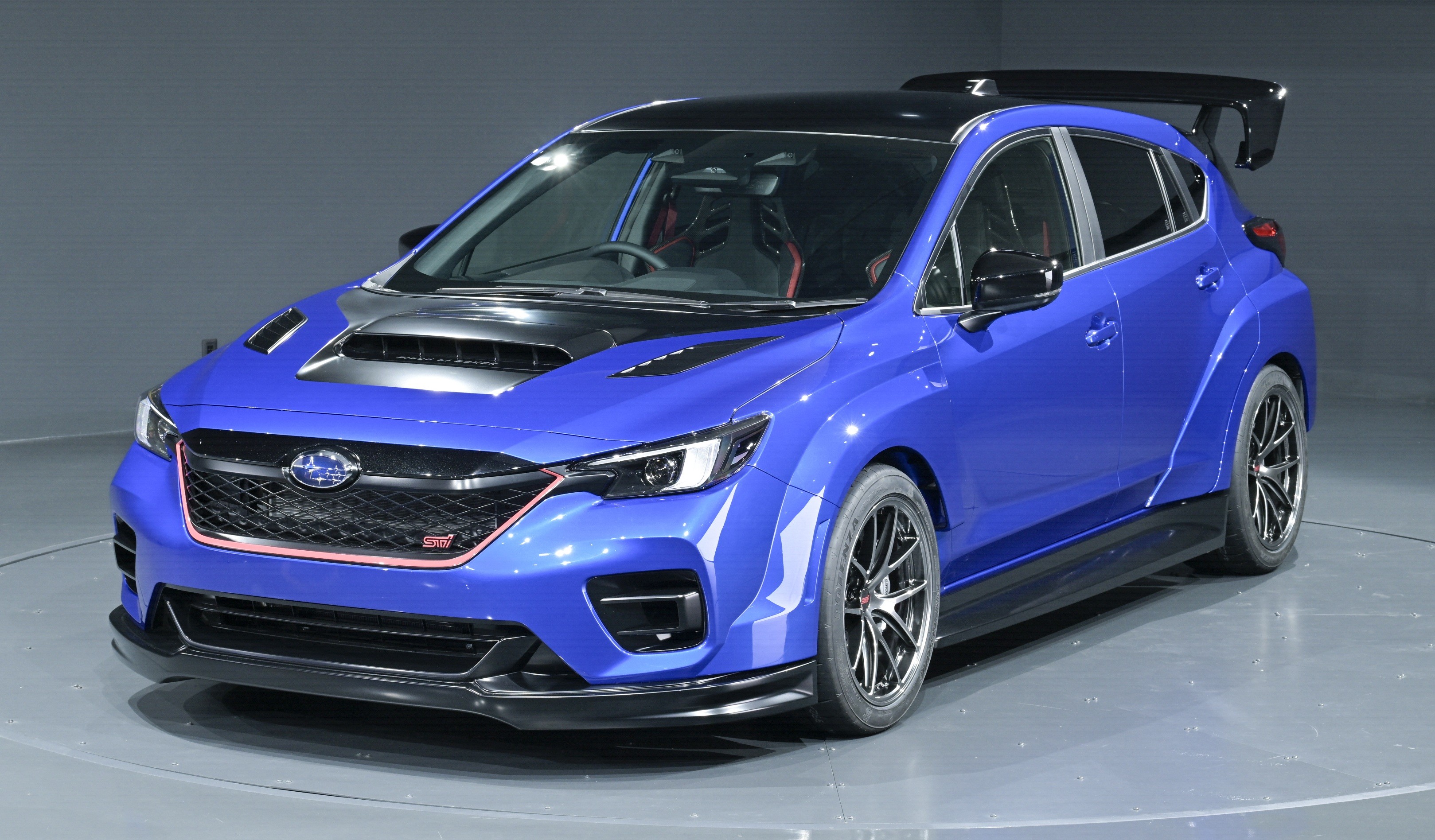 Carro esportivo azul met&aacute;lico com cap&ocirc; preto e entradas de ar, grade frontal preta com contorno vermelho e emblema STI, rodas escuras e aerof&oacute;lio traseiro, exibido em um est&uacute;dio com fundo cinza