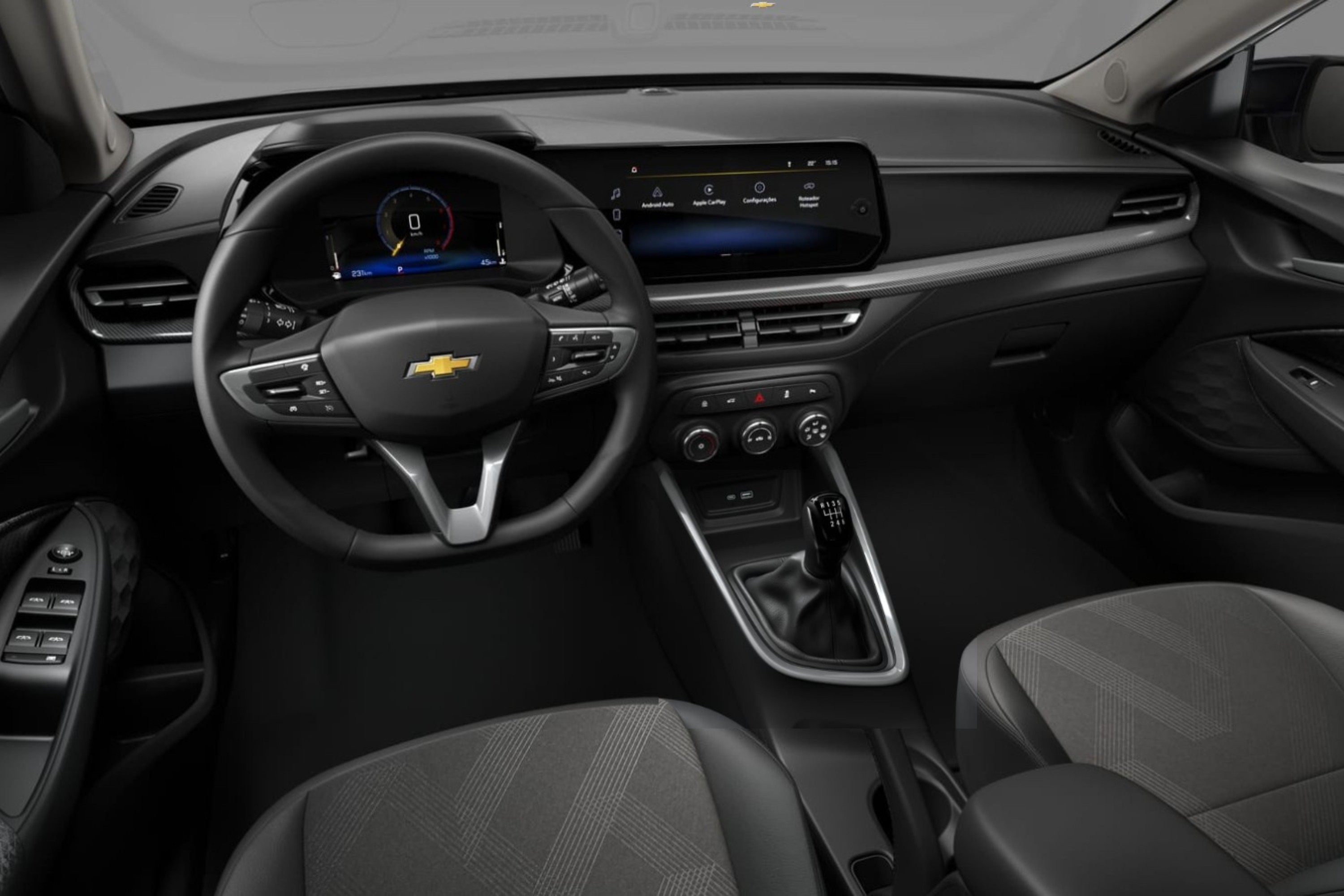 Interior de um carro moderno, com volante multifuncional preto e logo dourado da Chevrolet. Painel digital exibe veloc&iacute;metro e tela multim&iacute;dia sens&iacute;vel ao toque. C&acirc;mbio manual de cinco marchas e bancos de tecido cinza escuro com detalhes geom&eacute;tricos. O ambiente &eacute; predominantemente preto e cinza, com detalhes prateados no painel e console central