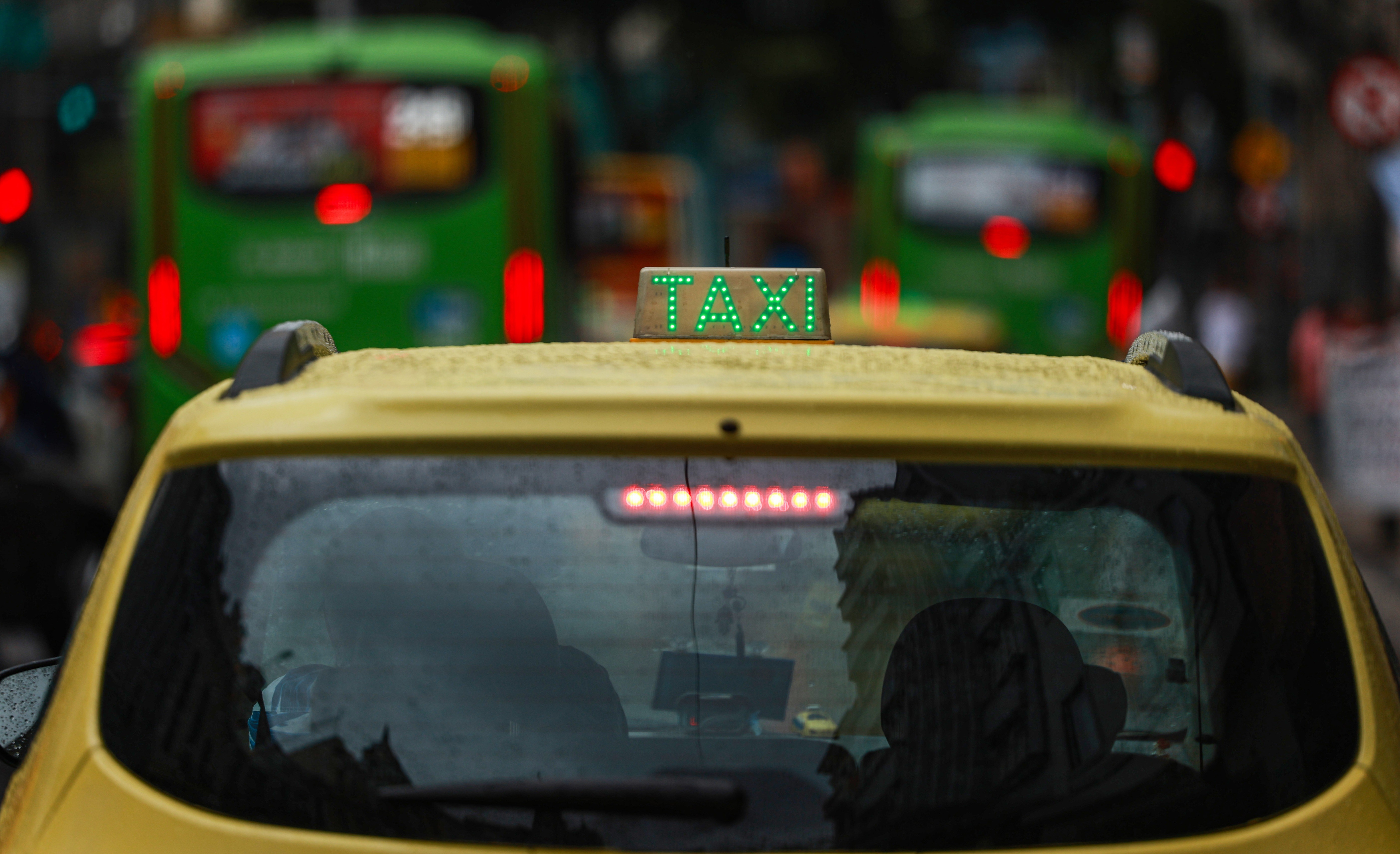 Traseira de um t&aacute;xi amarelo com o letreiro TAXI em verde luminoso no teto, em meio ao tr&acirc;nsito urbano. Gotas de chuva cobrem o vidro traseiro e o teto do carro. Ao fundo, &ocirc;nibus verdes e luzes vermelhas de freio est&atilde;o desfocados, indicando movimento em uma rua movimentada