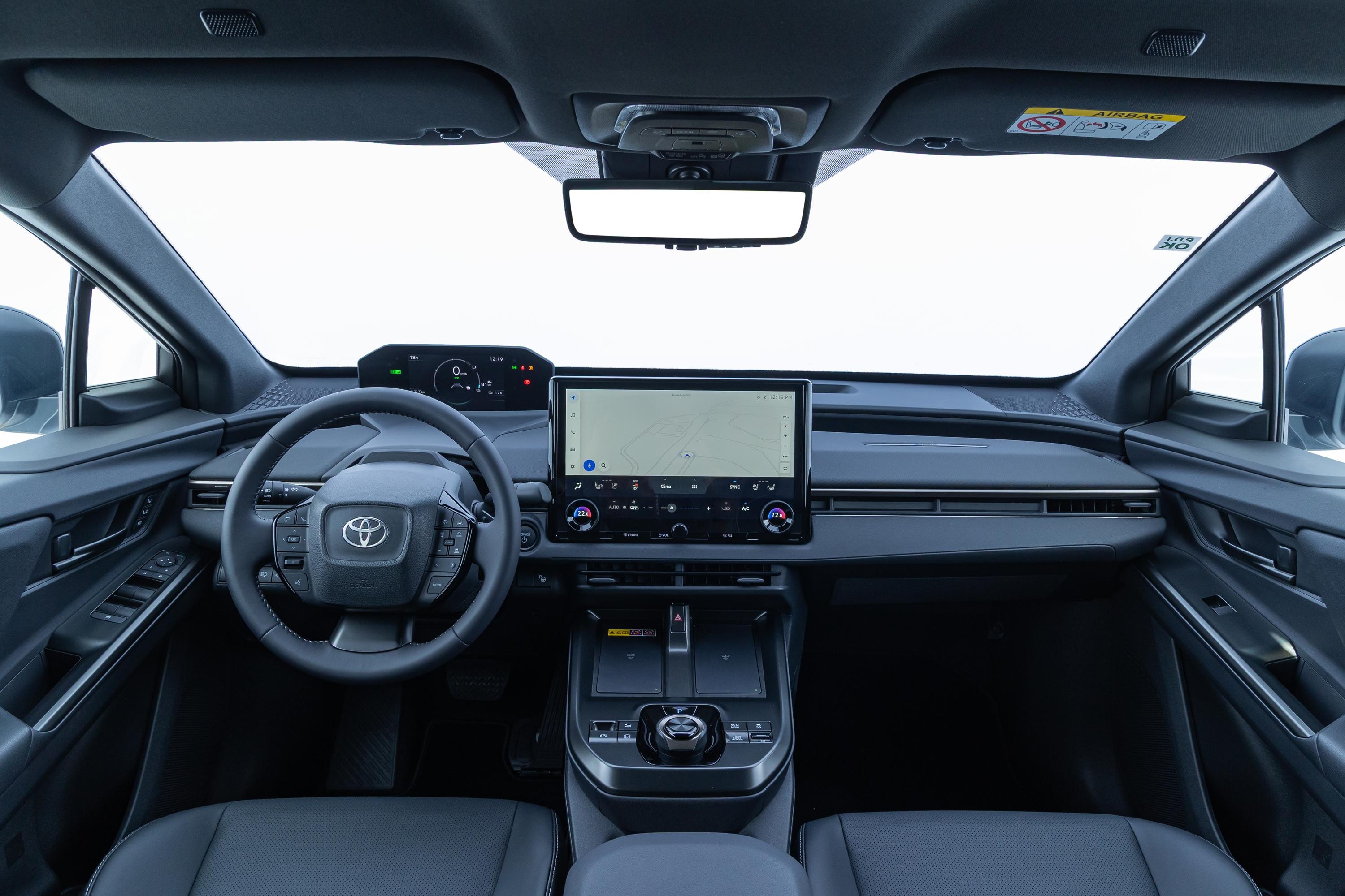 Interior de um carro moderno, com painel escuro e volante multifuncional com o logo da Toyota. H&aacute; uma tela multim&iacute;dia grande no centro e um painel de instrumentos digital acima do volante. O para-brisa e as janelas laterais mostram um fundo branco, sugerindo um est&uacute;dio ou c&eacute;u claro. Os bancos s&atilde;o de couro escuro e o console central possui bot&otilde;es e porta-objetos