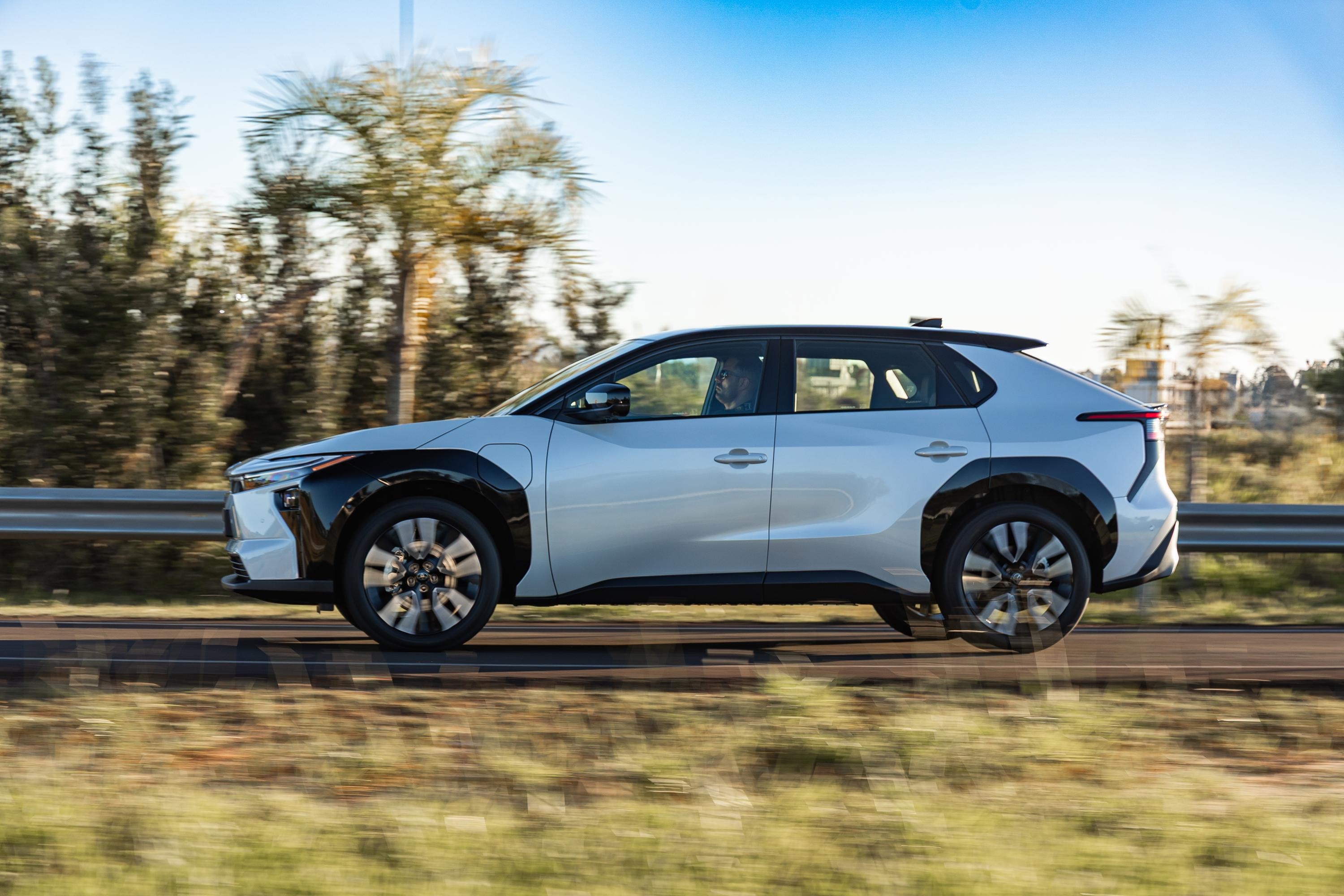 SUV el&eacute;trico branco e preto em movimento, visto de perfil, com fundo desfocado de &aacute;rvores e c&eacute;u azul